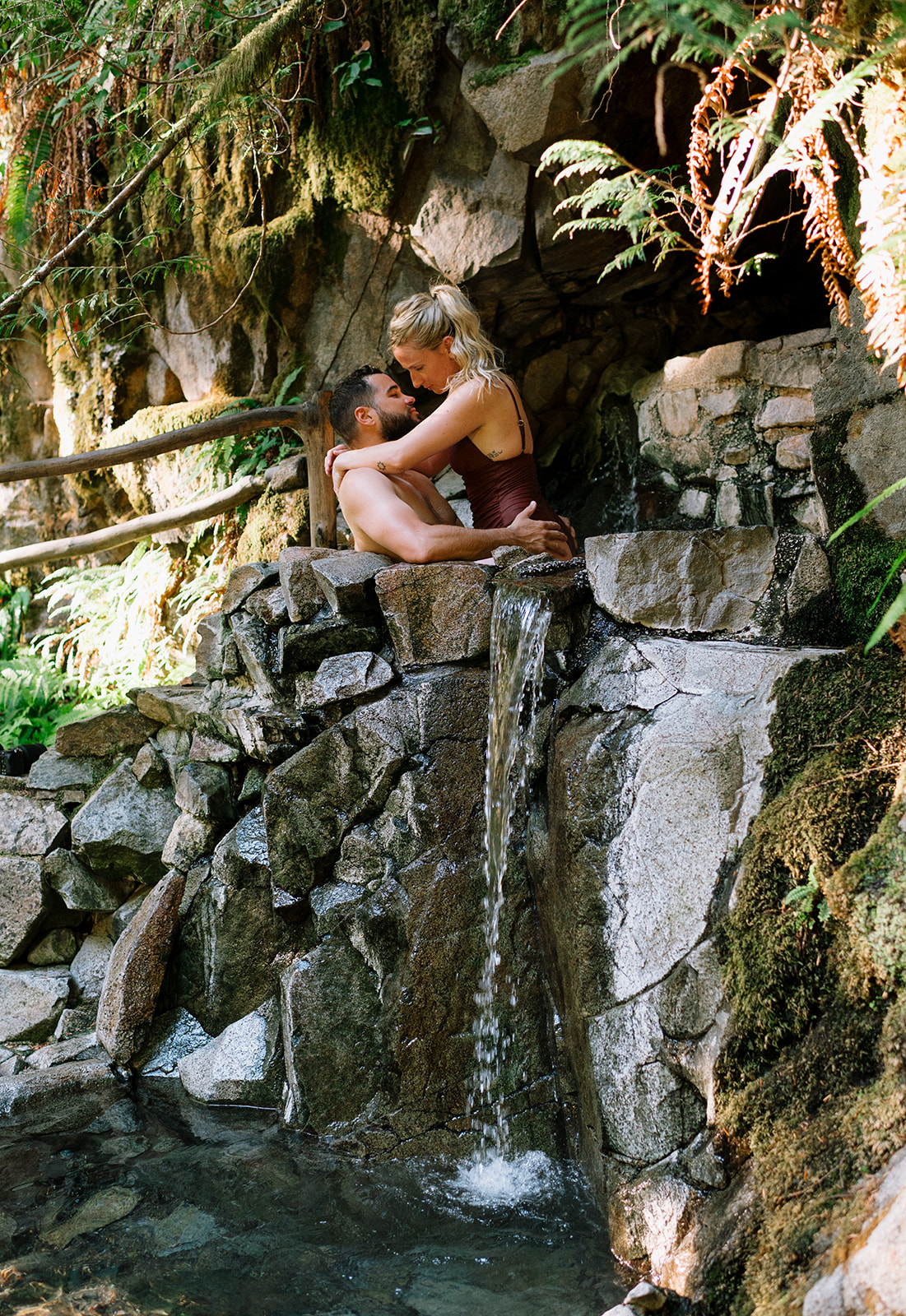 R and M embracing in a hot spring with a small waterfall in the background.