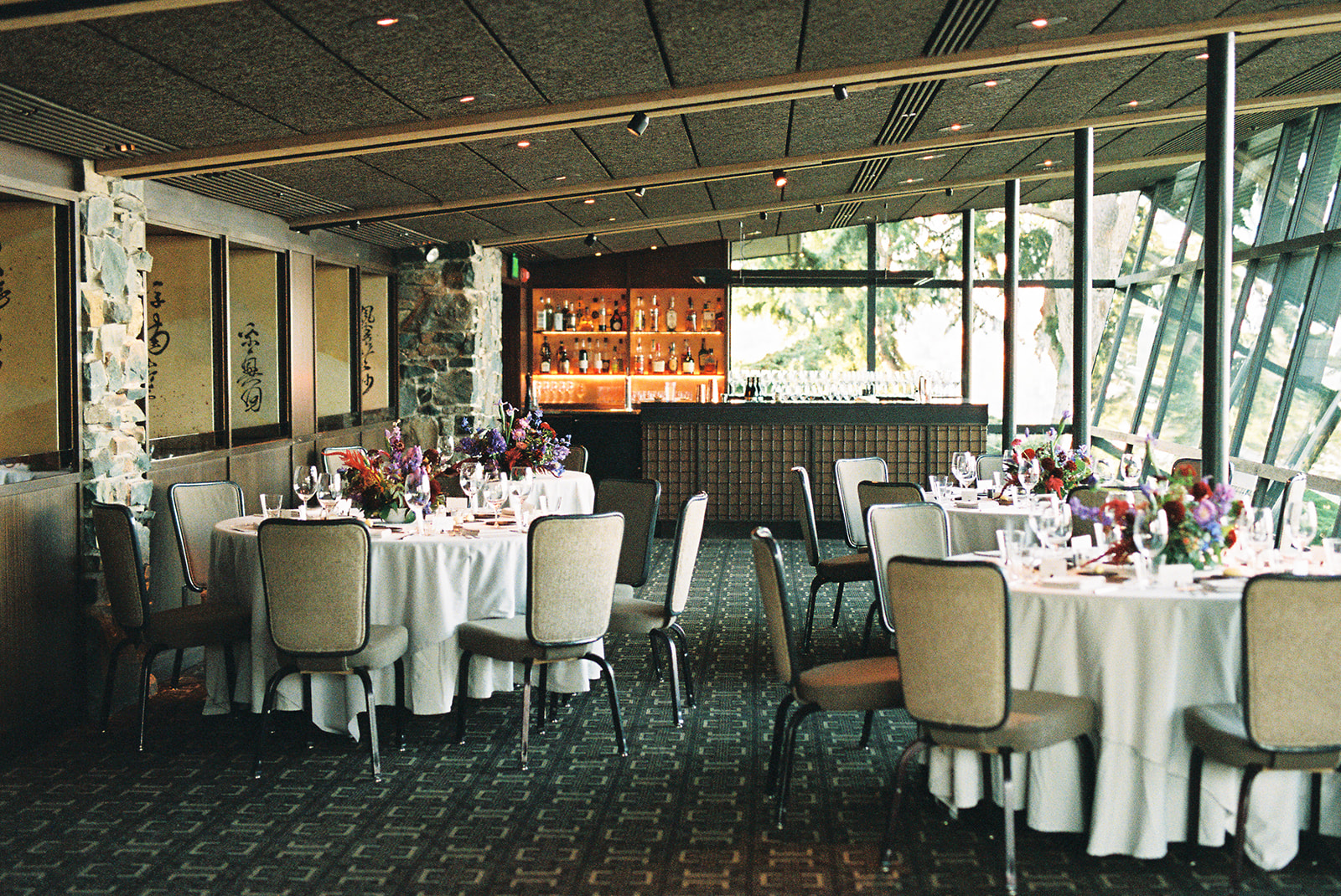 Elegant dining room at Canlis with round tables set with white linens, floral centerpieces, and a well-stocked bar in the background.