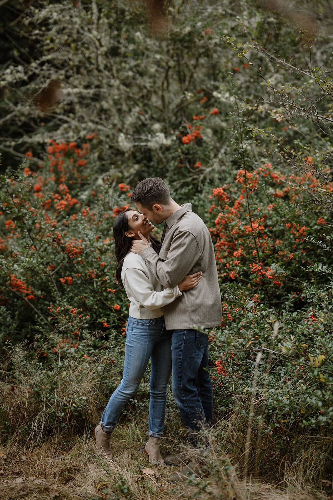 Couple kisses in front of vibrant red holly bushes surrounded by fall foliage at Discovery Park.