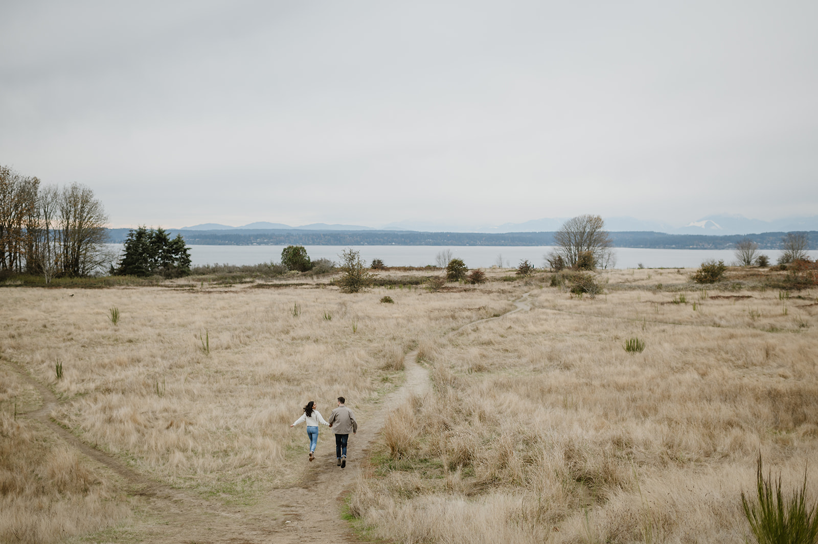 Couple walks hand in hand through an open field with Puget Sound in the background.