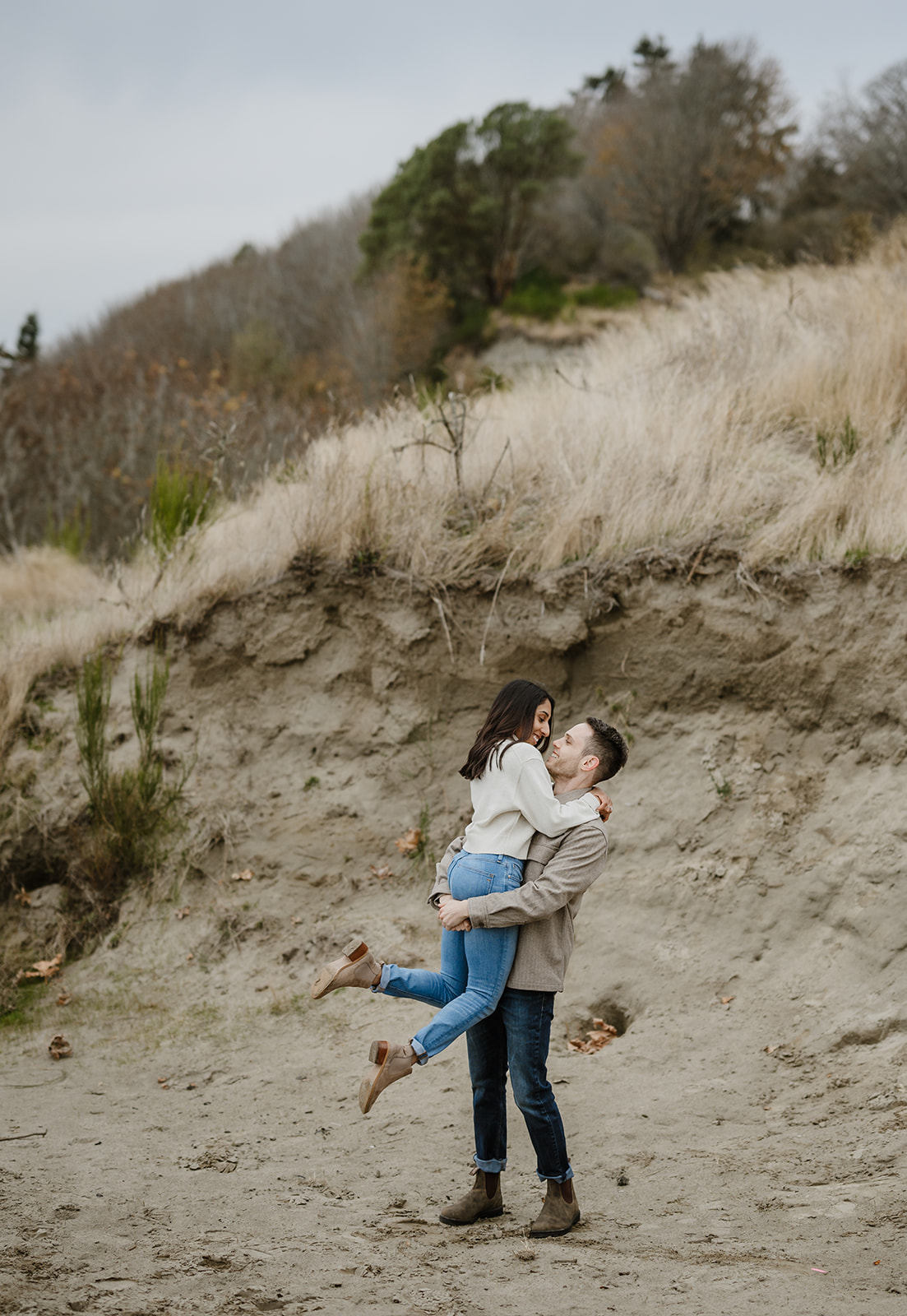 Man lifts his partner in a playful moment on a sandy hillside during a fall engagement session.