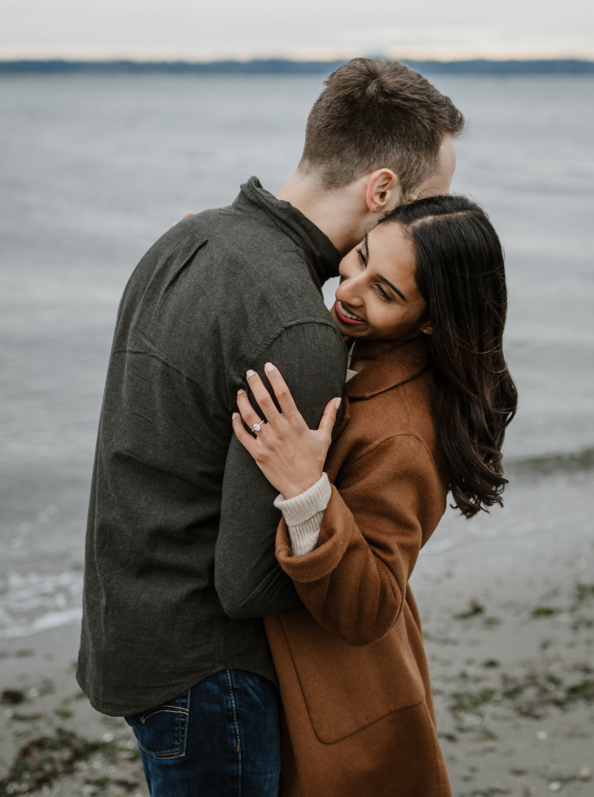 A couple embraces joyfully by the shore, smiling as they share a moment of affection on a misty day at the beach.