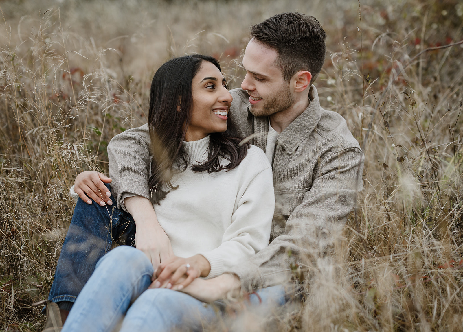 Couple sitting together in a field of tall grasses, gazing at each other with joy and affection.