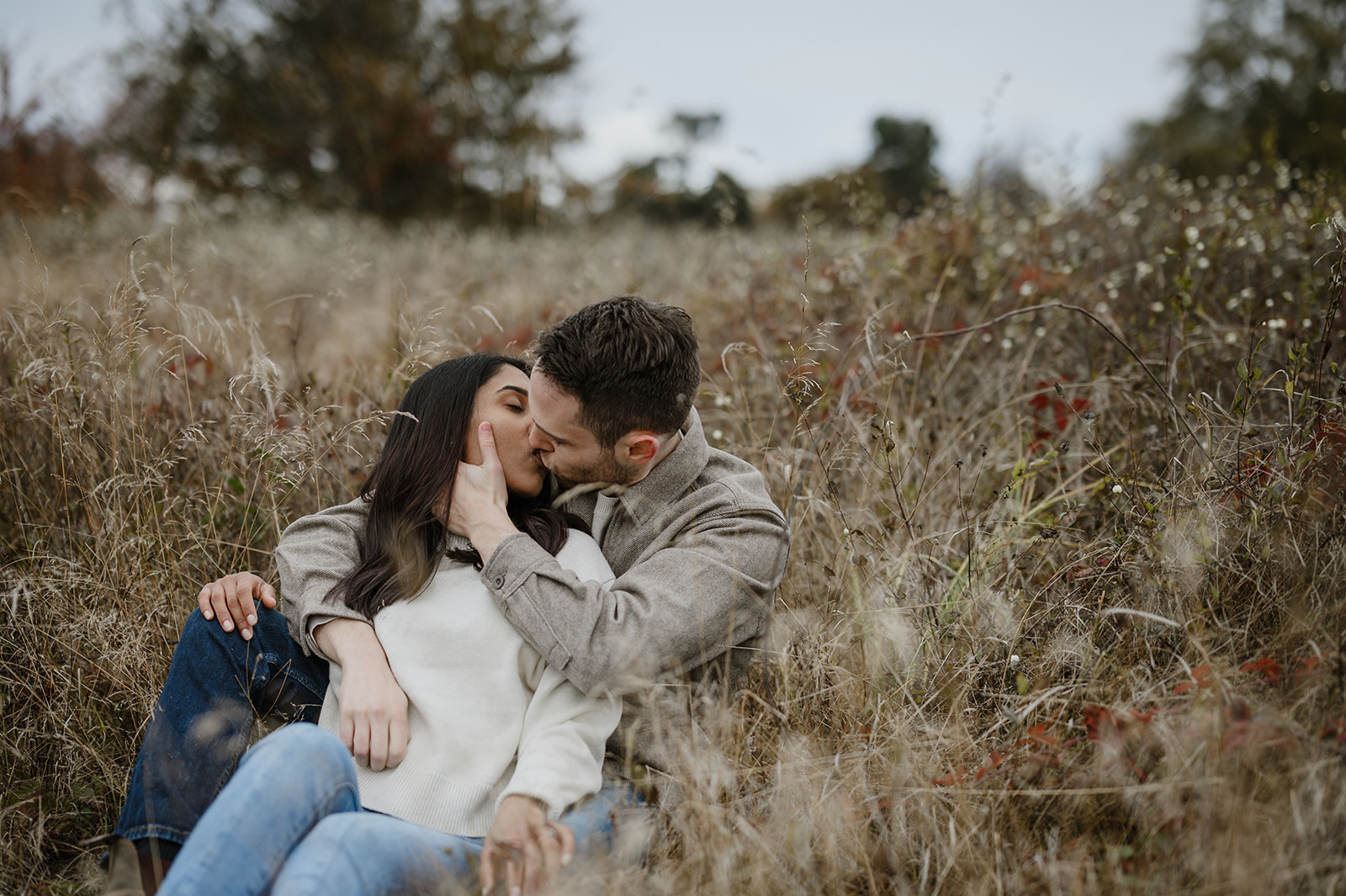 Couple kissing while sitting in a field of tall grasses, surrounded by autumn foliage.