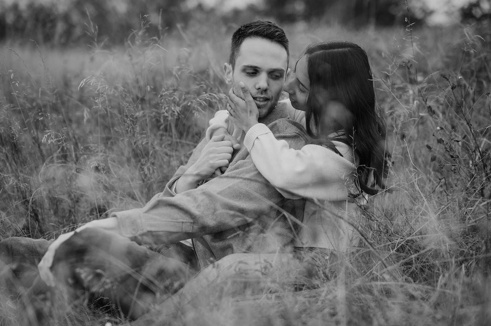 A couple sits close in a field, gently embracing as they share a kiss amidst tall grasses.