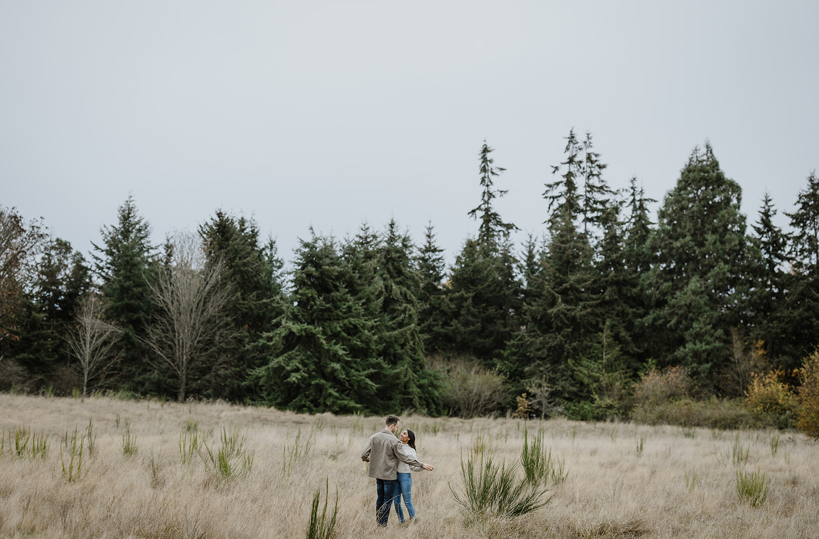 Couple holding each other closely in an open grassy field, surrounded by towering trees, enjoying the peacefulness of nature.