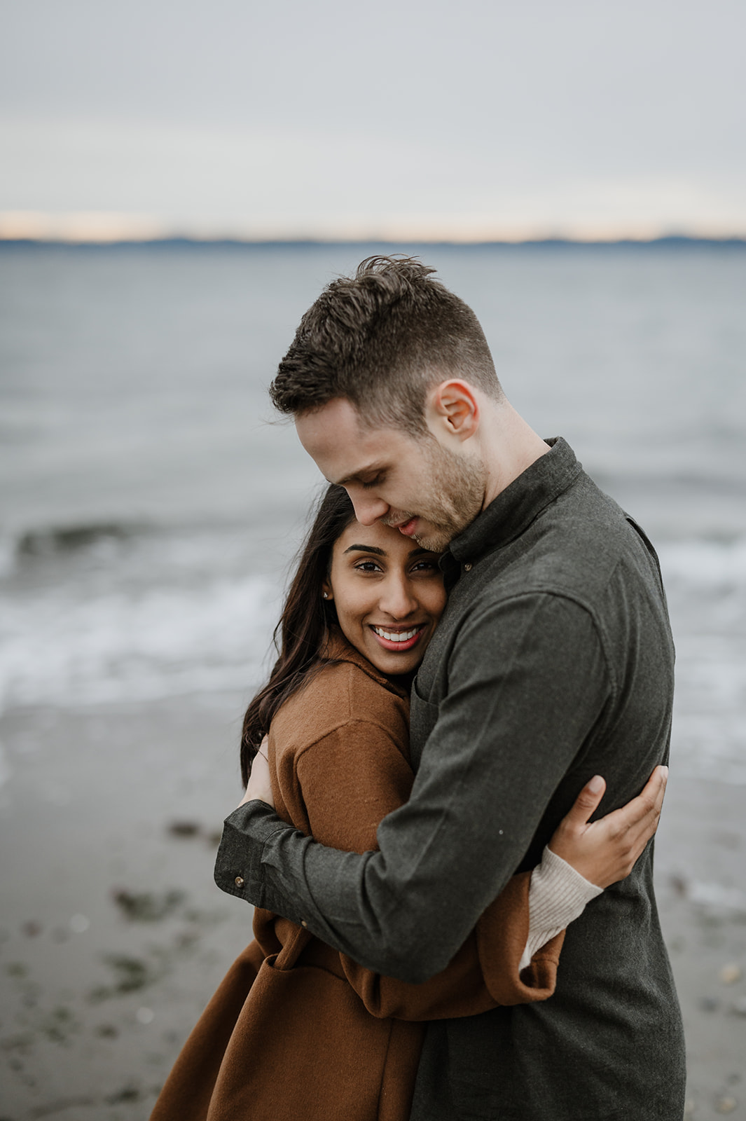 Couple hugging on a Seattle beach with the woman smiling brightly at the camera.