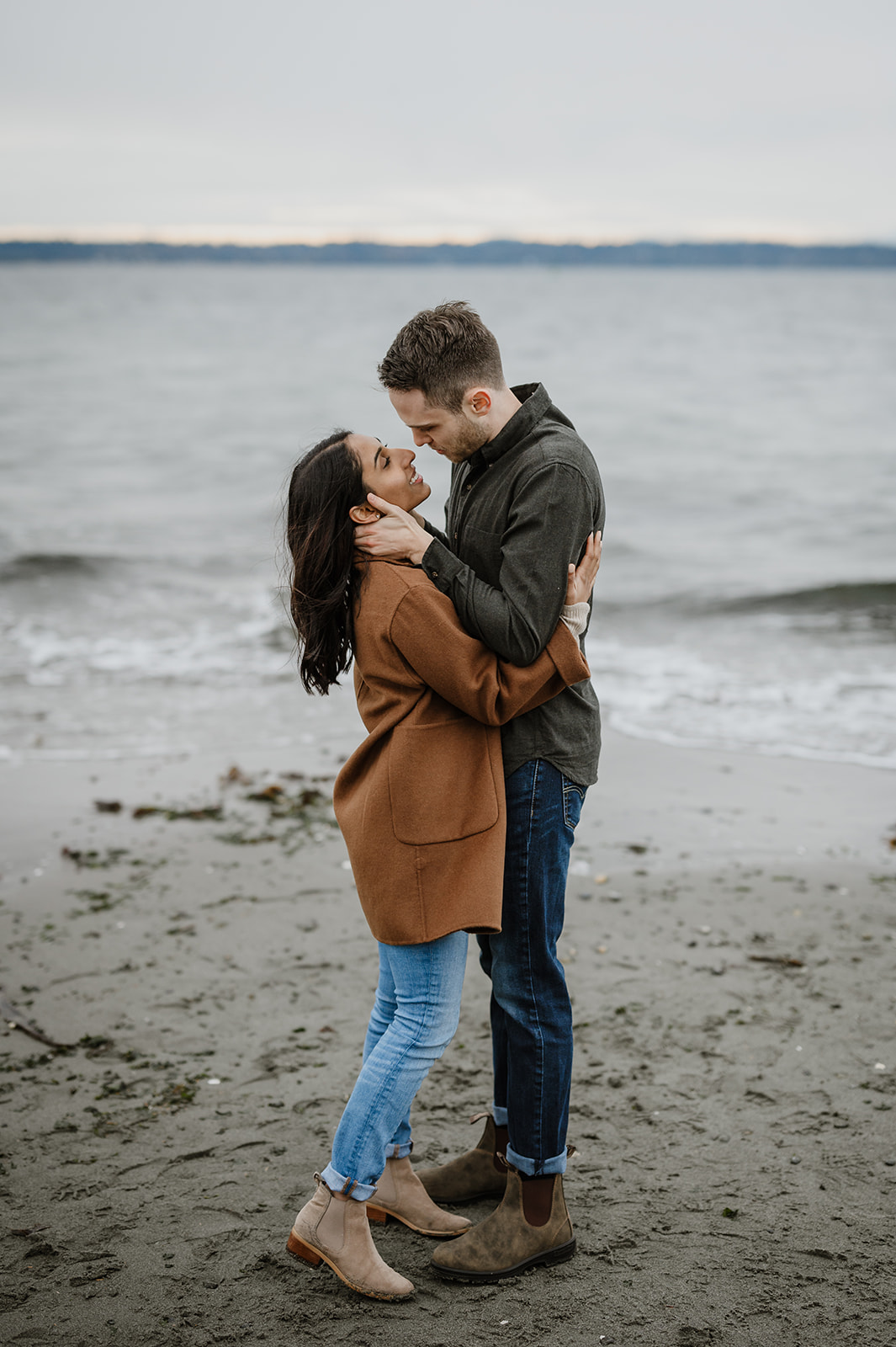 A couple stands close on the sandy shore, gently holding each other as they gaze into each other's eyes at Discovery Park.