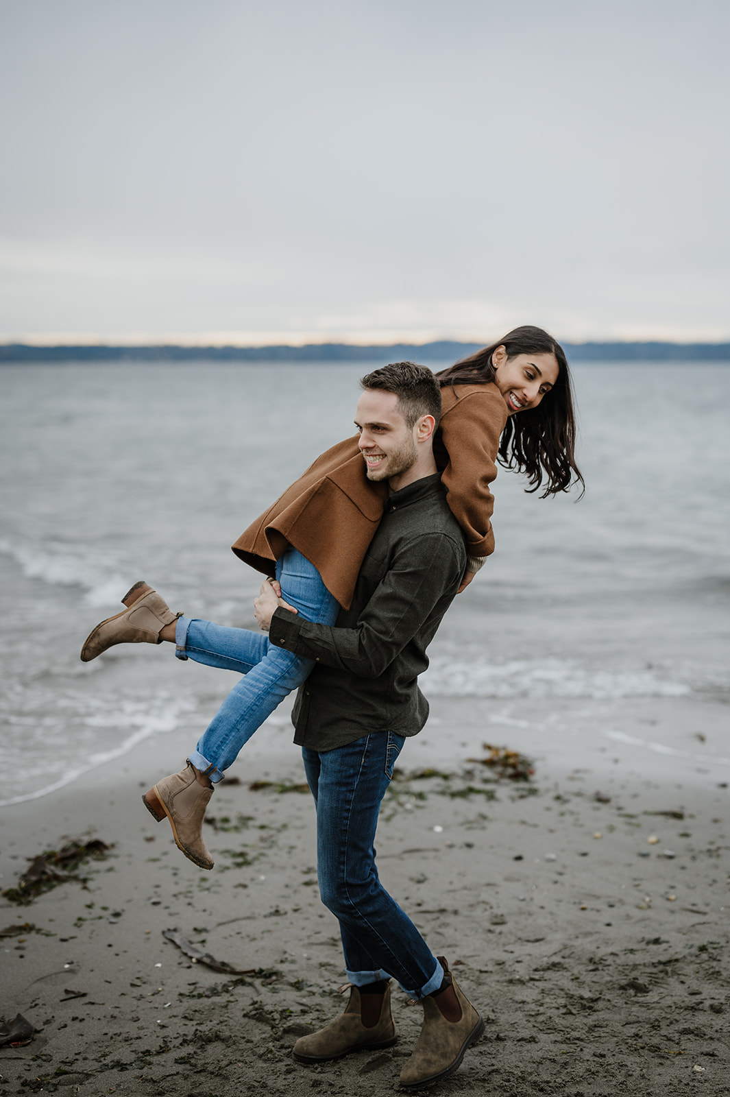 A man lifts his partner playfully as they both smile, enjoying a lighthearted moment by the water's edge at the beach.