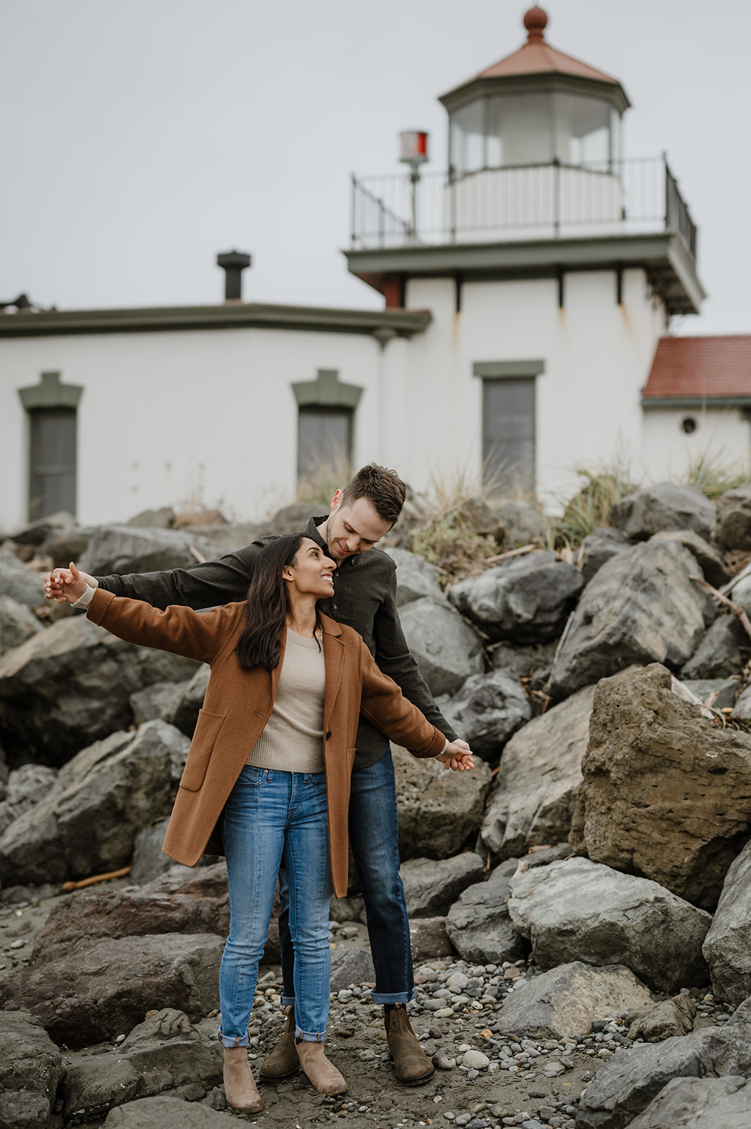 Couple standing in front of a lighthouse with the woman stretching her arms open in joy.