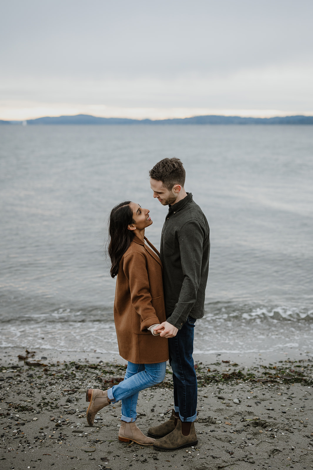 Couple holding hands and smiling at each other while standing on the beach.