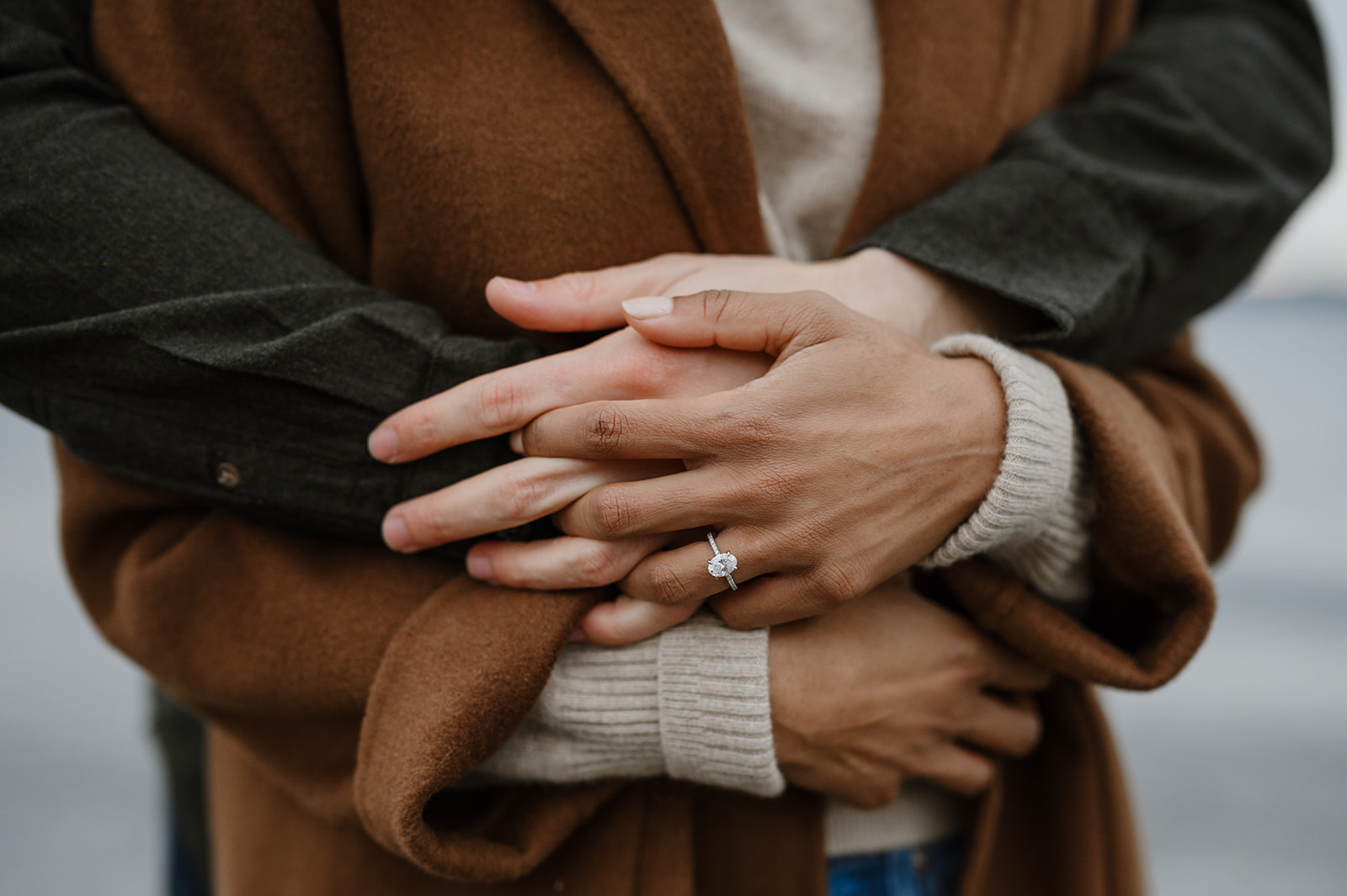 Close-up of a couple's hands, the woman's engagement ring visible as they stand by the beach.