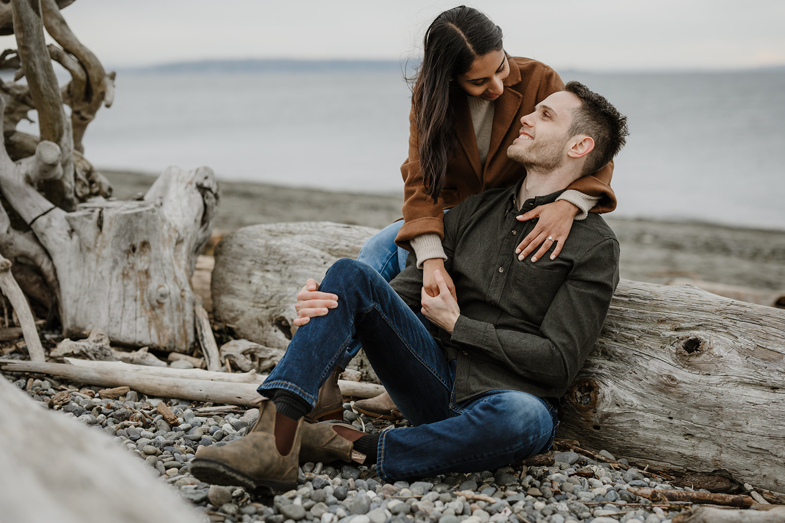 Couple sitting on driftwood along a rocky beach, holding hands and gazing into each other’s eyes.