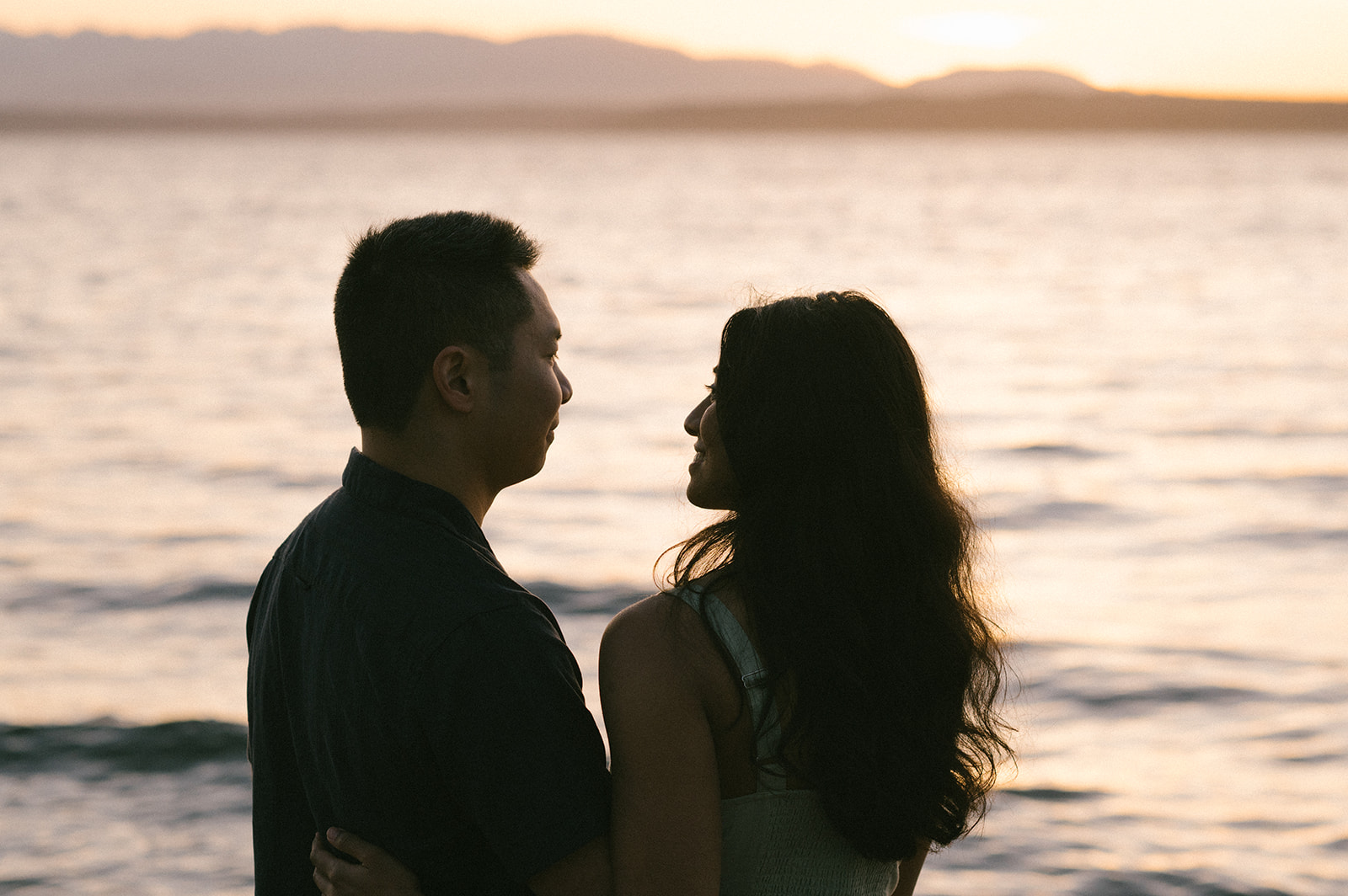A couple silhouetted against the sunset, looking into each other's eyes by the water.