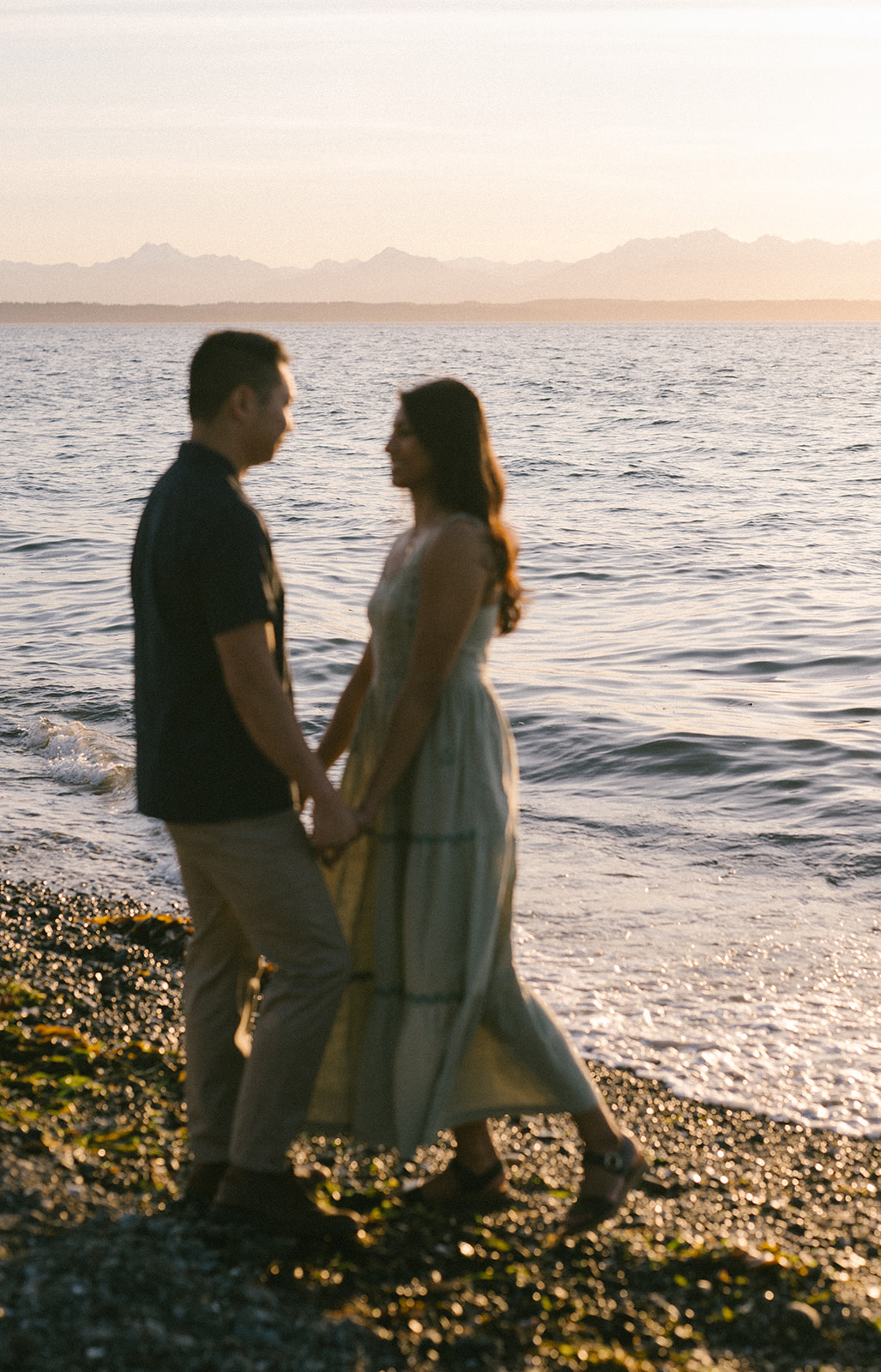 A couple holding hands, silhouetted against the sunset by the water.