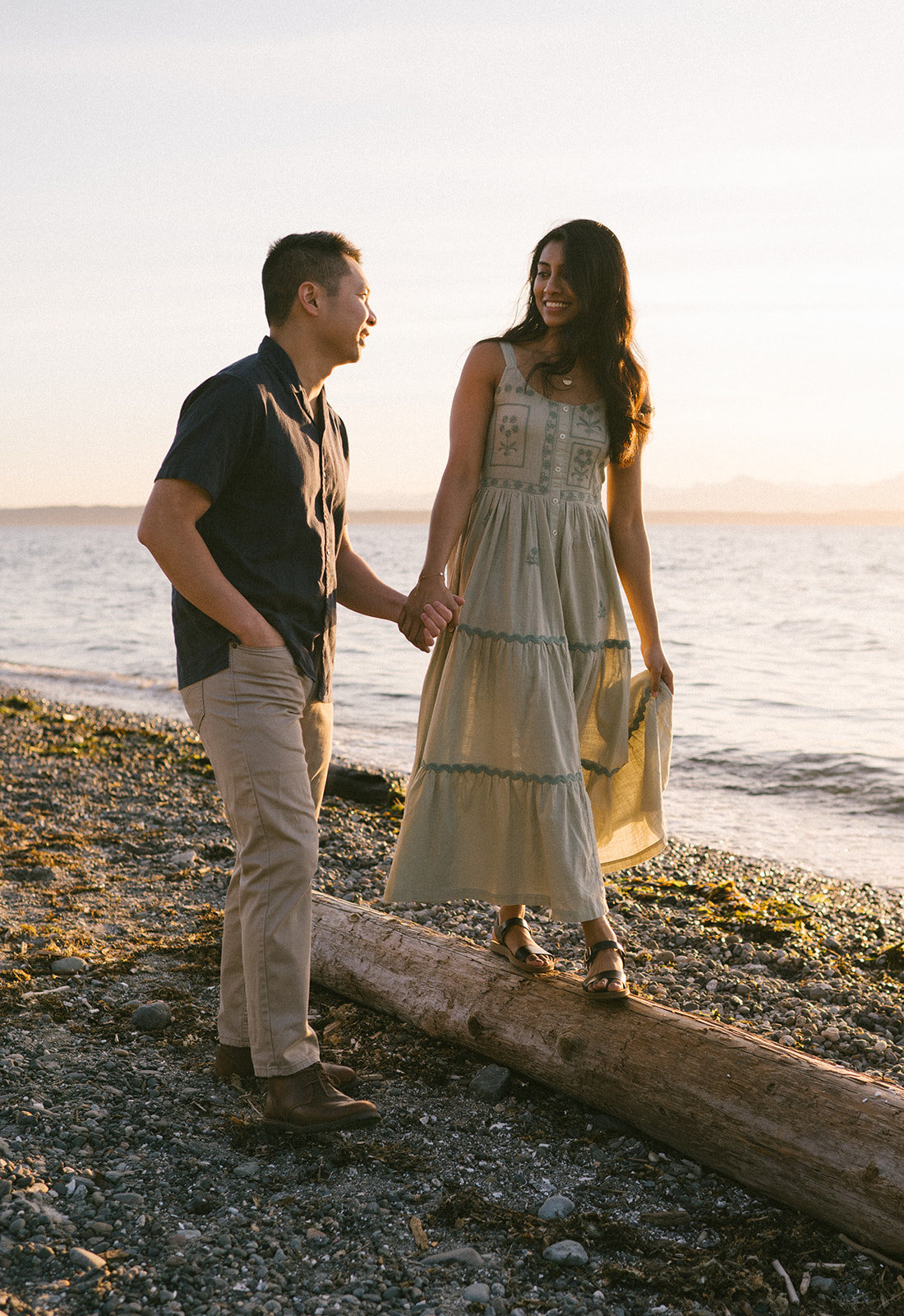 A couple walking along a log on the beach, holding hands at sunset.