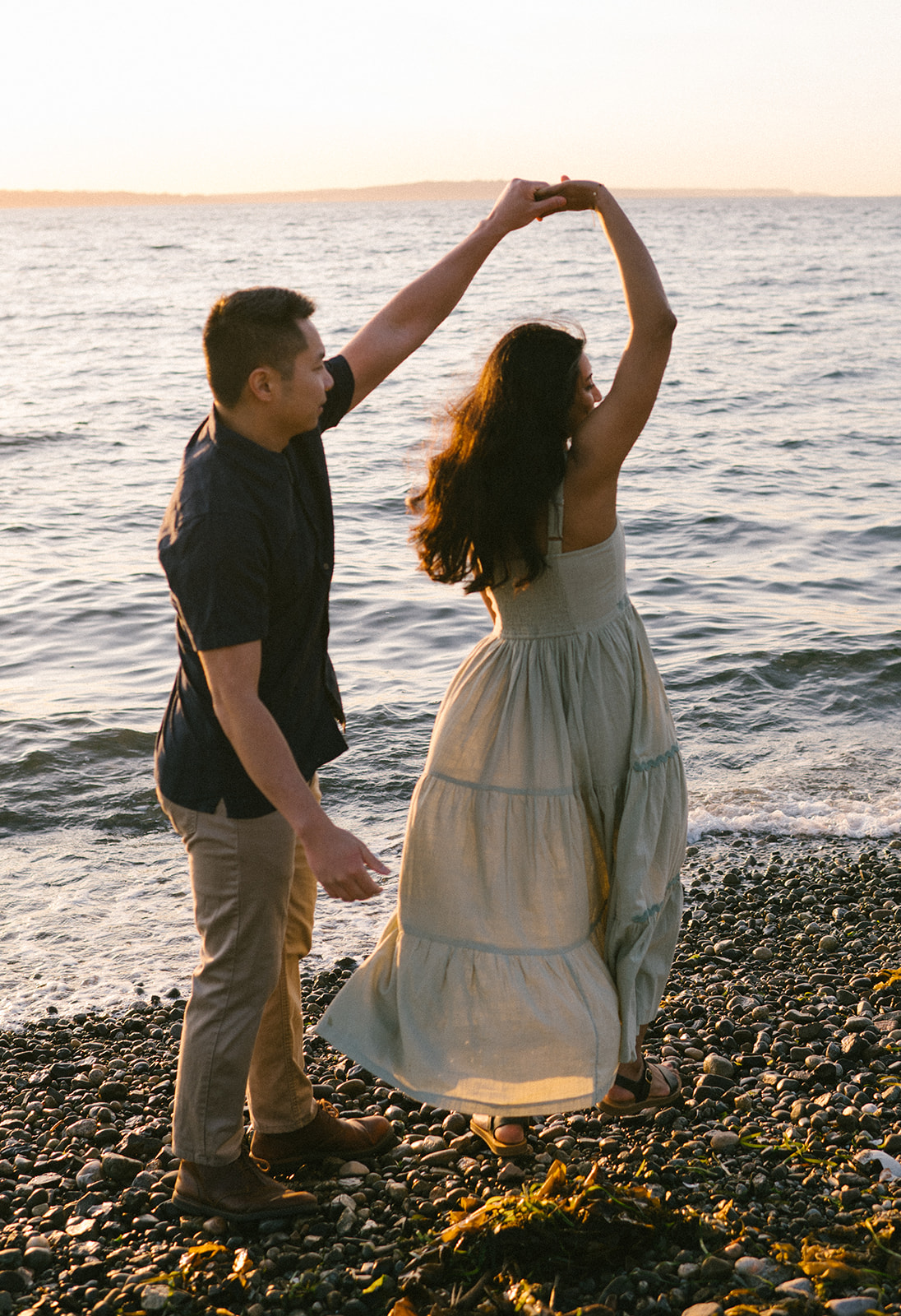 A couple dancing by the water at sunset, enjoying a lighthearted moment.