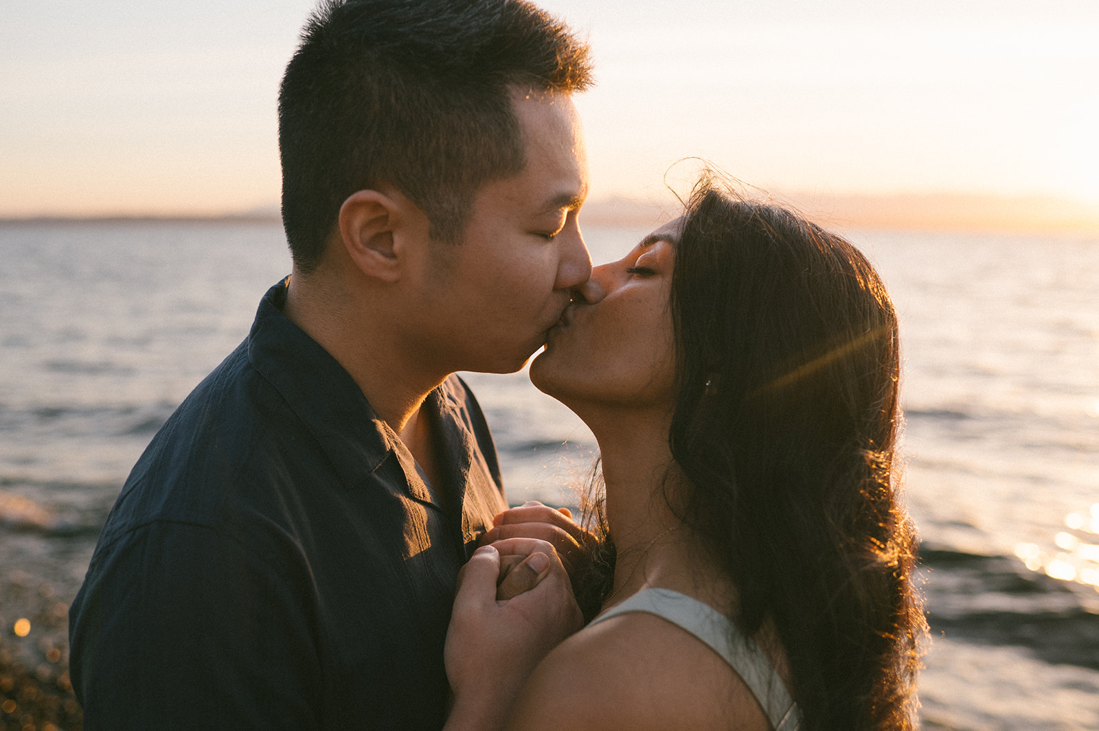 A close-up of a couple kissing and holding hands, focusing on their connection. The sun setting behind them. 