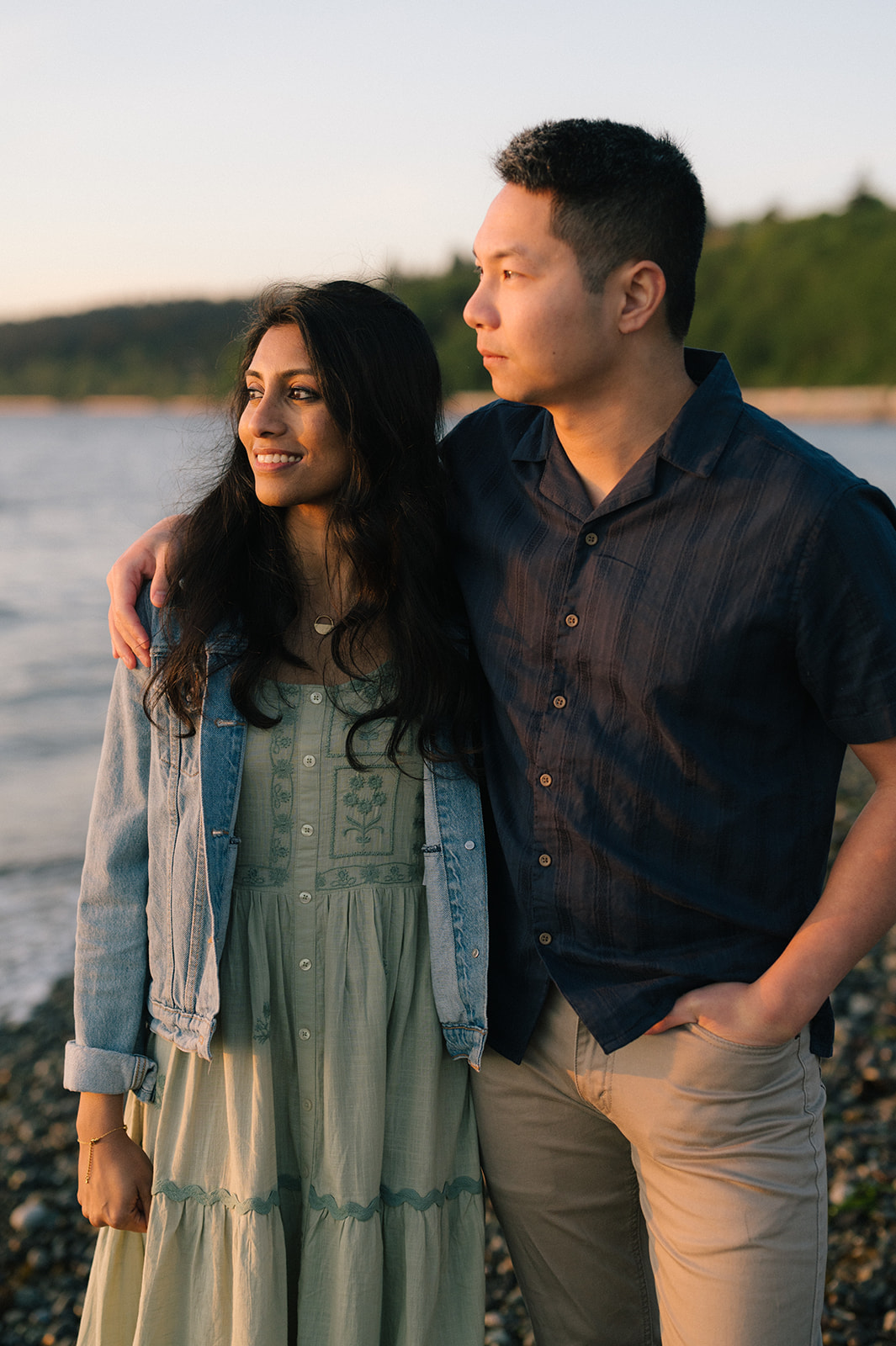 A couple standing by the water, with arms around each other, looking at the sunset.