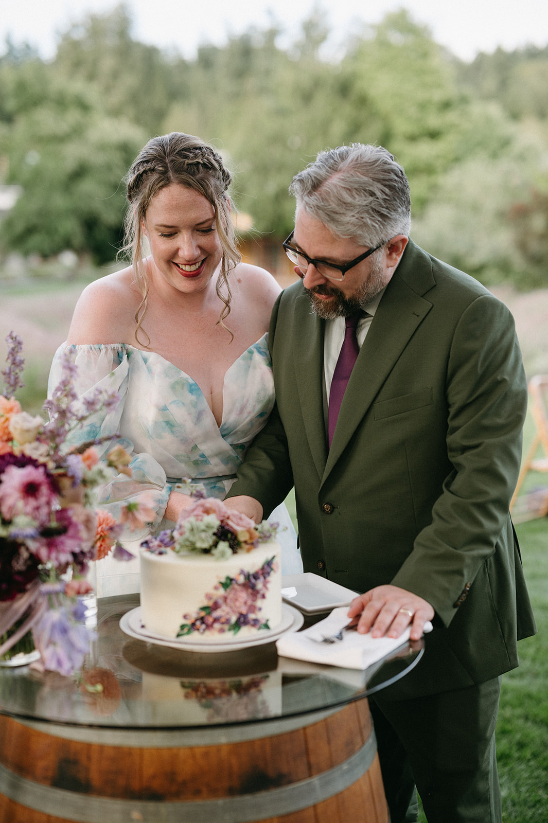 Couple smiling and cutting their wedding cake together outdoors.