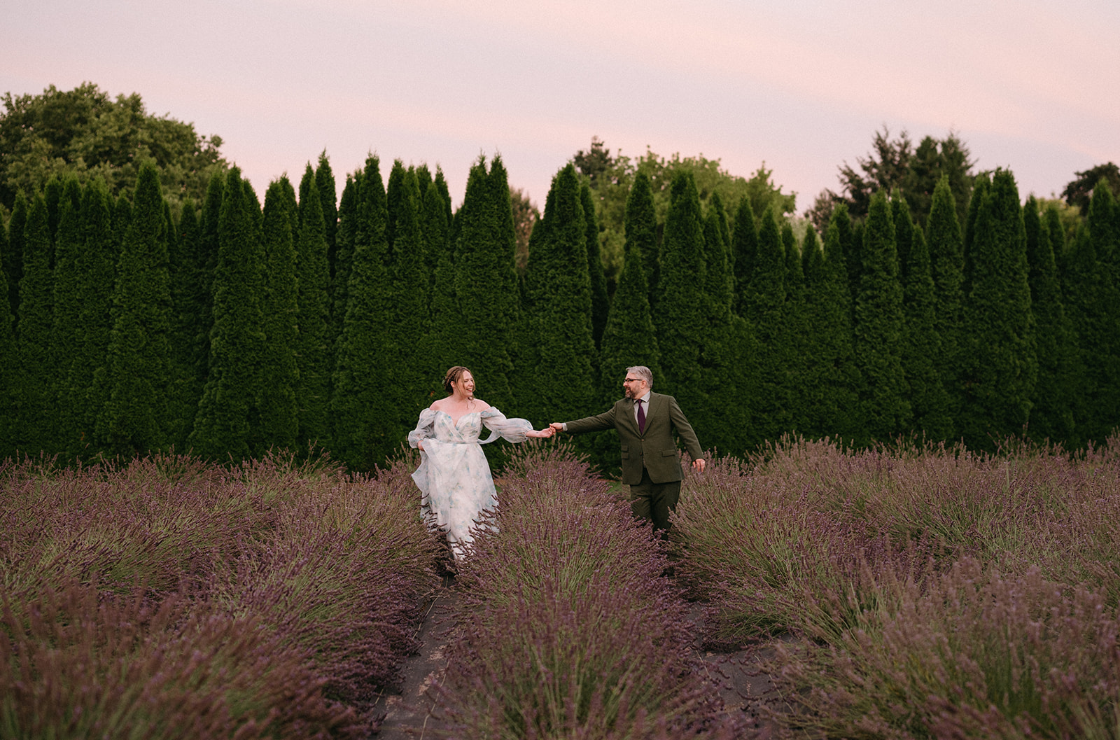 Couple running hand in hand through rows of lavender with a sunset sky overhead.