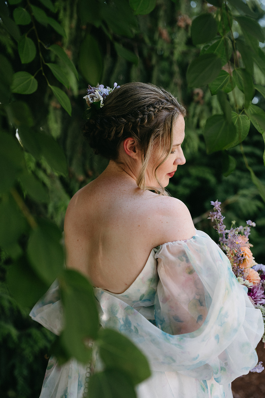 Bride facing away, framed by leaves, with floral braid and off-the-shoulder gown