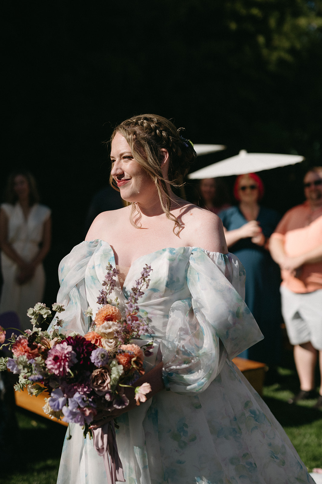 Bride smiling with bouquet during outdoor ceremony, guests in background