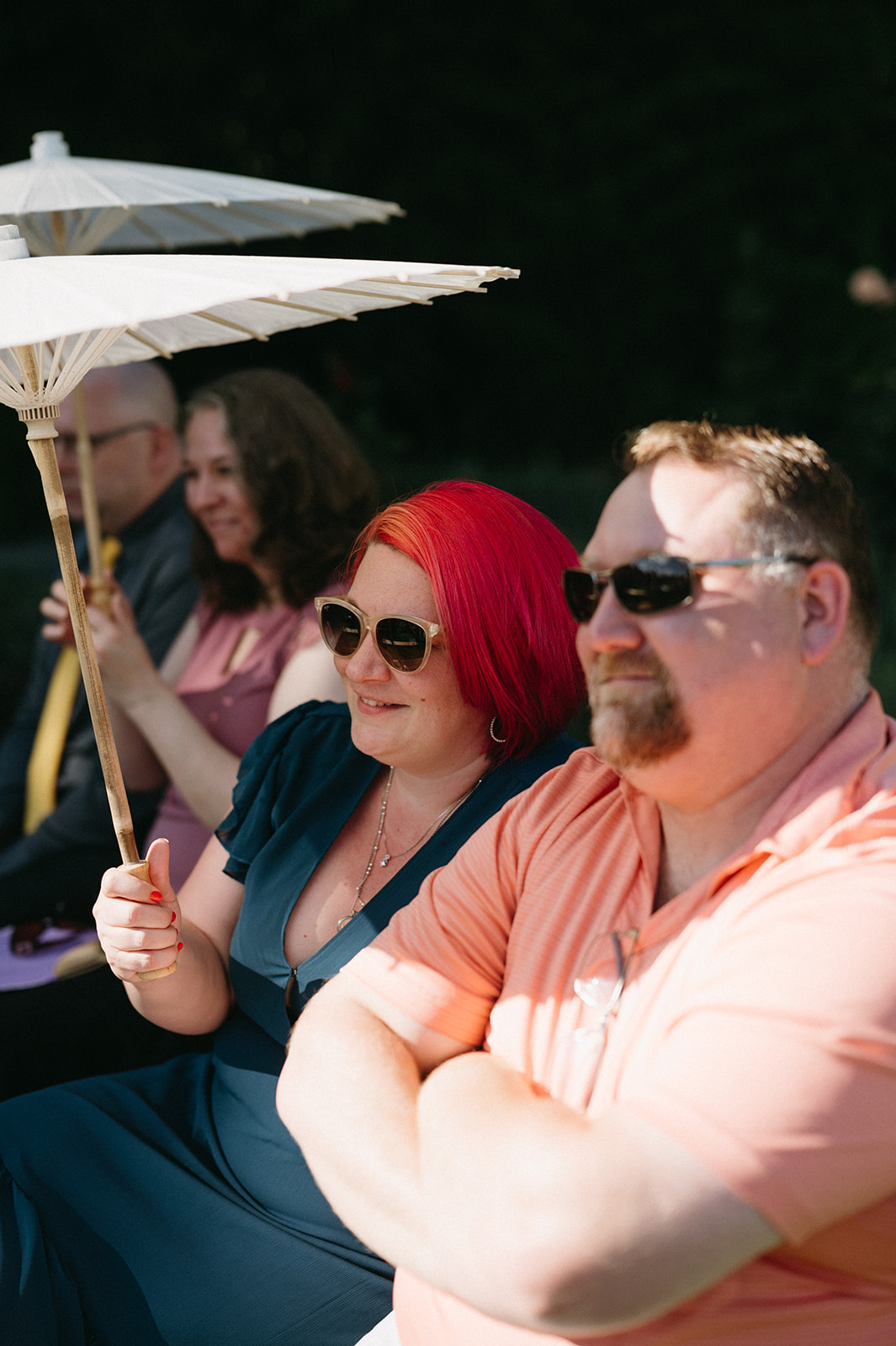 Wedding guest holding parasol, seated in sunlight during outdoor ceremony