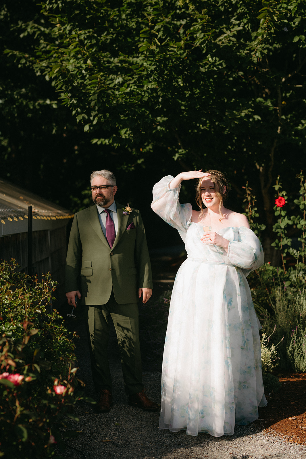 Couple standing near a garden path, bride shielding eyes from sun, groom holding a glass.