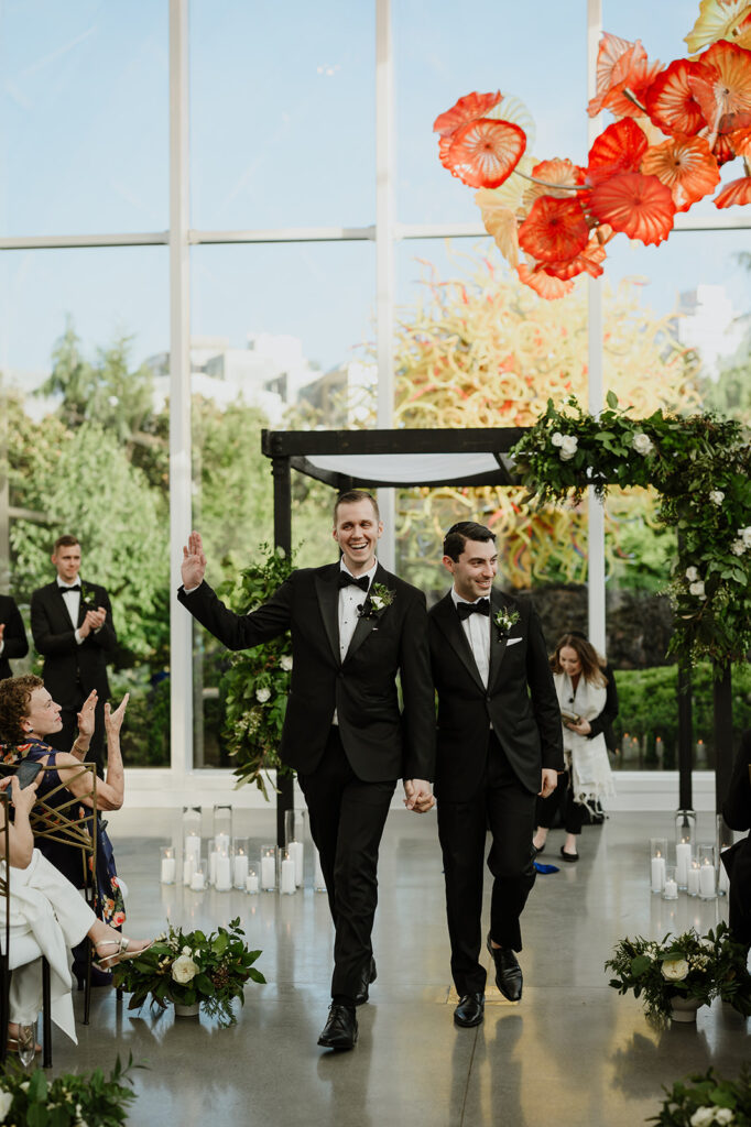 Two grooms walking down the aisle as newlyweds at a Chihuly Garden and Glass wedding ceremony