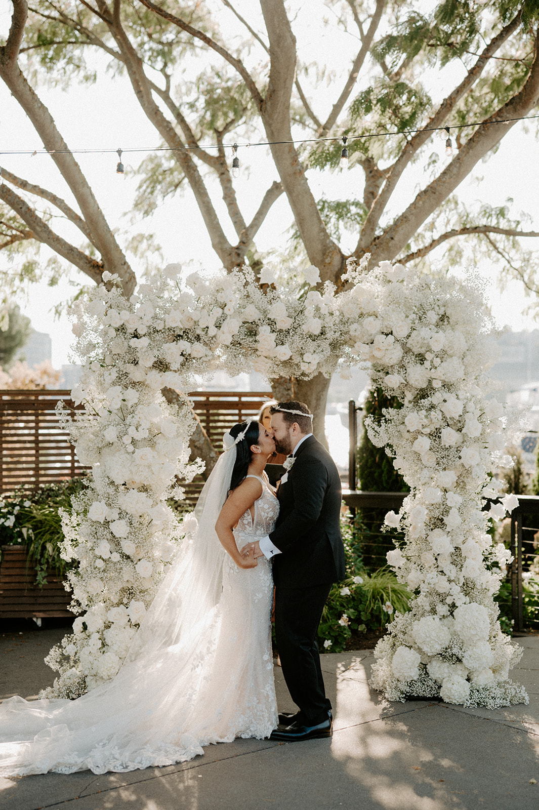 The bride and groom share their first kiss under a large floral arch at their outdoor wedding ceremony.