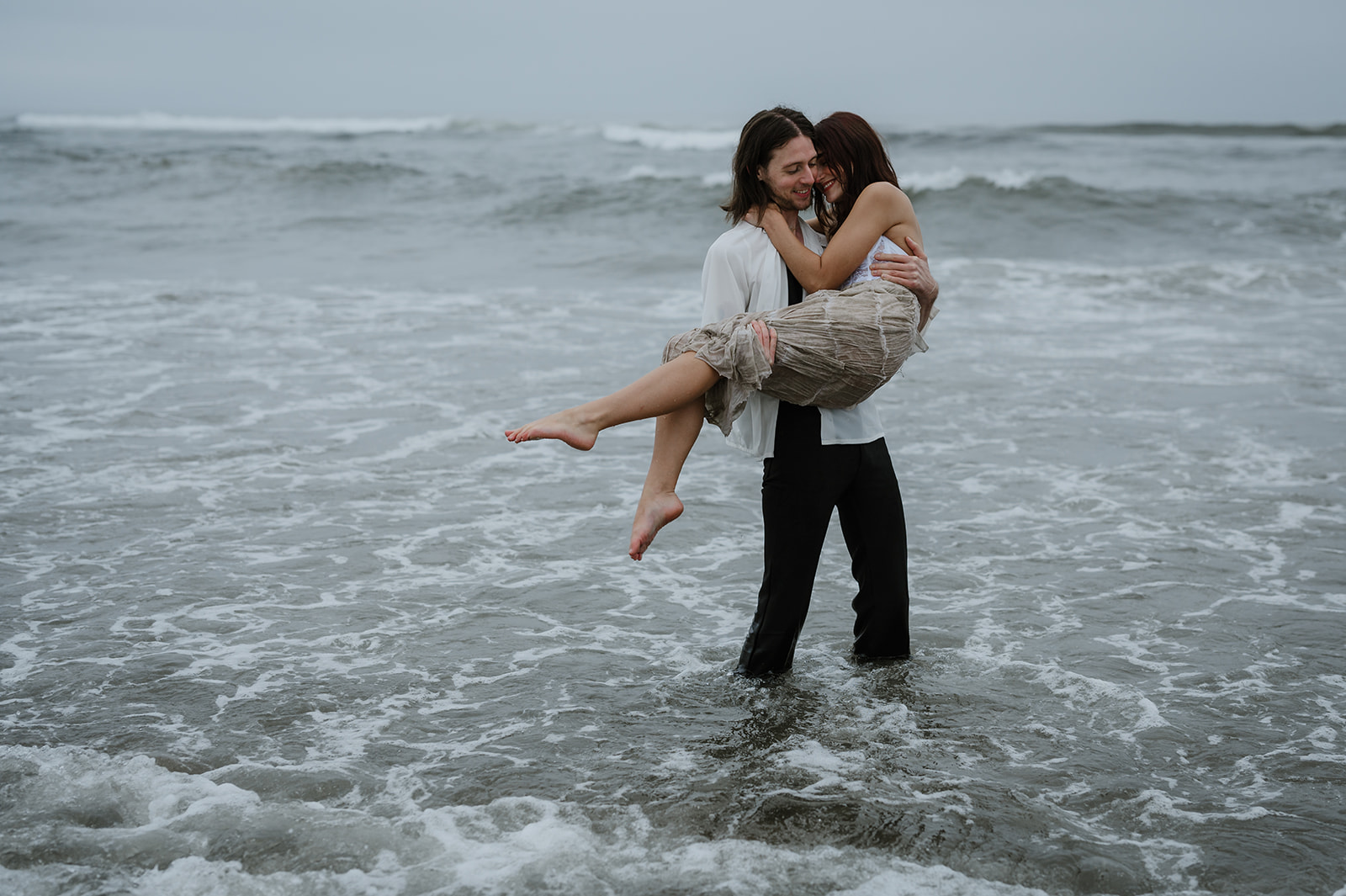 A groom carrying his bride in the ocean waves, both smiling under overcast skies.