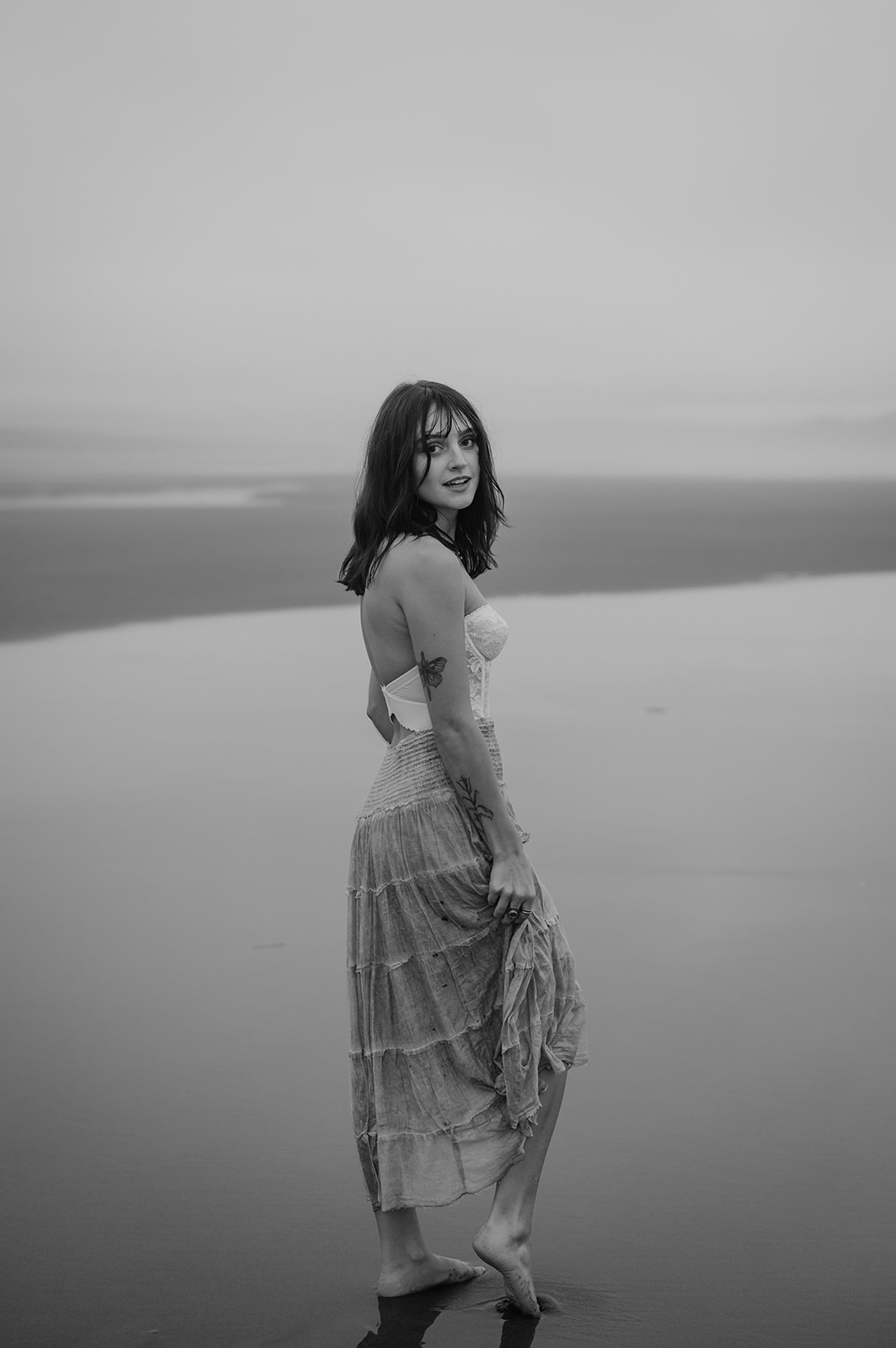 A bride walking barefoot on a misty beach, looking over her shoulder with a serene expression.