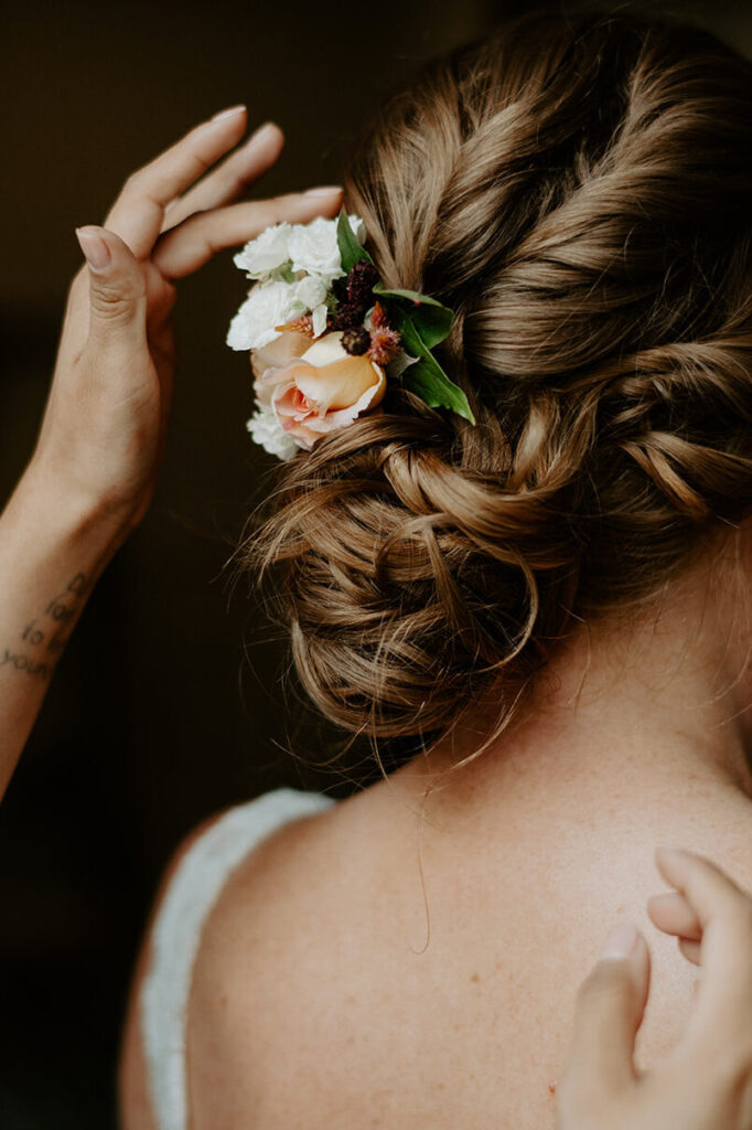 A close-up of a bride's intricately styled hair adorned with fresh flowers, as a hand adjusts the arrangement.