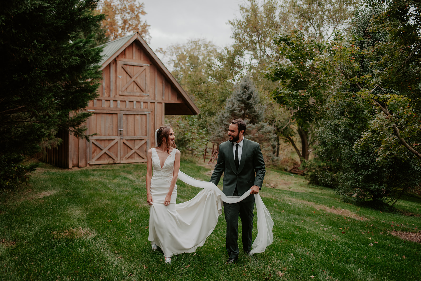 Bride and groom walking hand-in-hand near a rustic barn, with the bride’s veil flowing in the breeze.