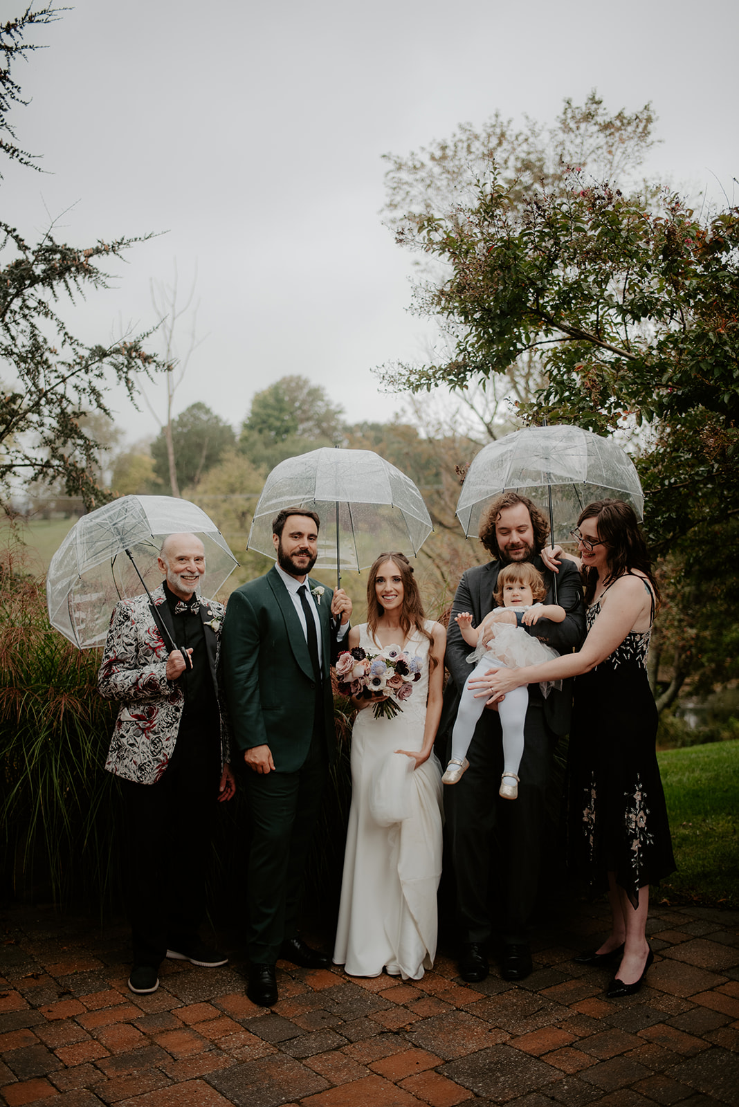 A wedding party standing together with clear umbrellas, surrounded by greenery and overcast skies.