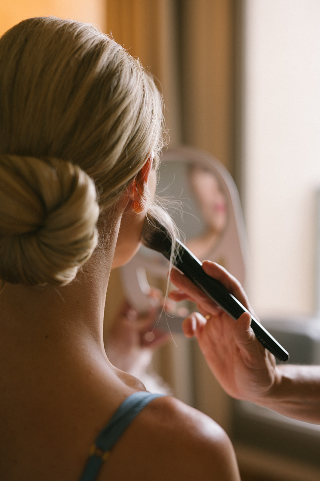 Close-up of bride’s low bun hairstyle as a makeup artist applies finishing touches near a window.
