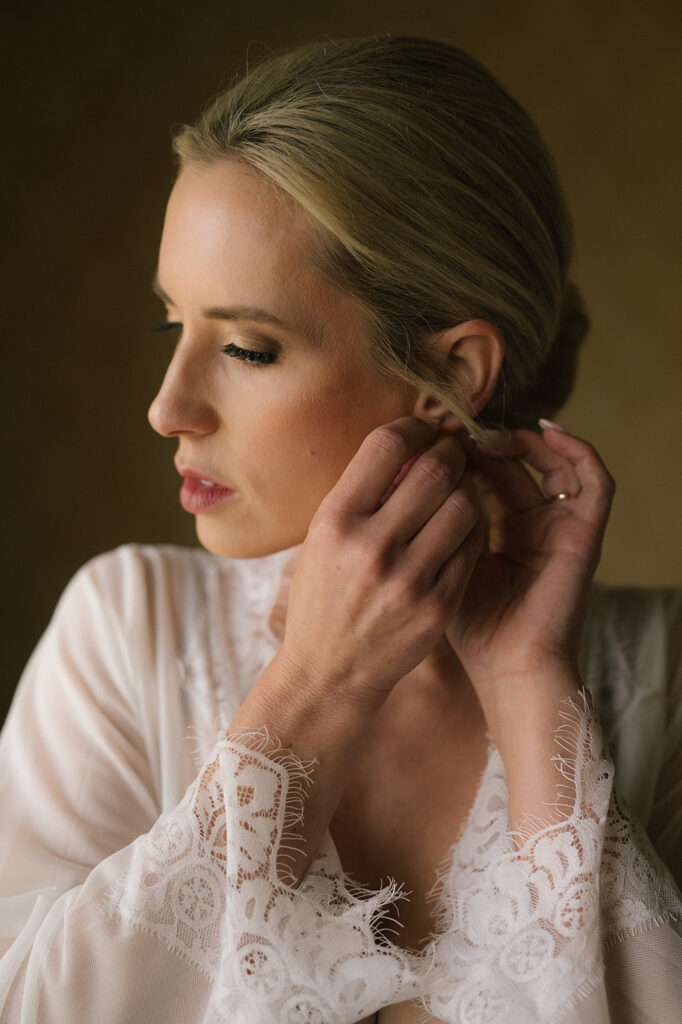 Bride putting on her earrings while wearing a lace robe in her suite at the Four Seasons Maui.
