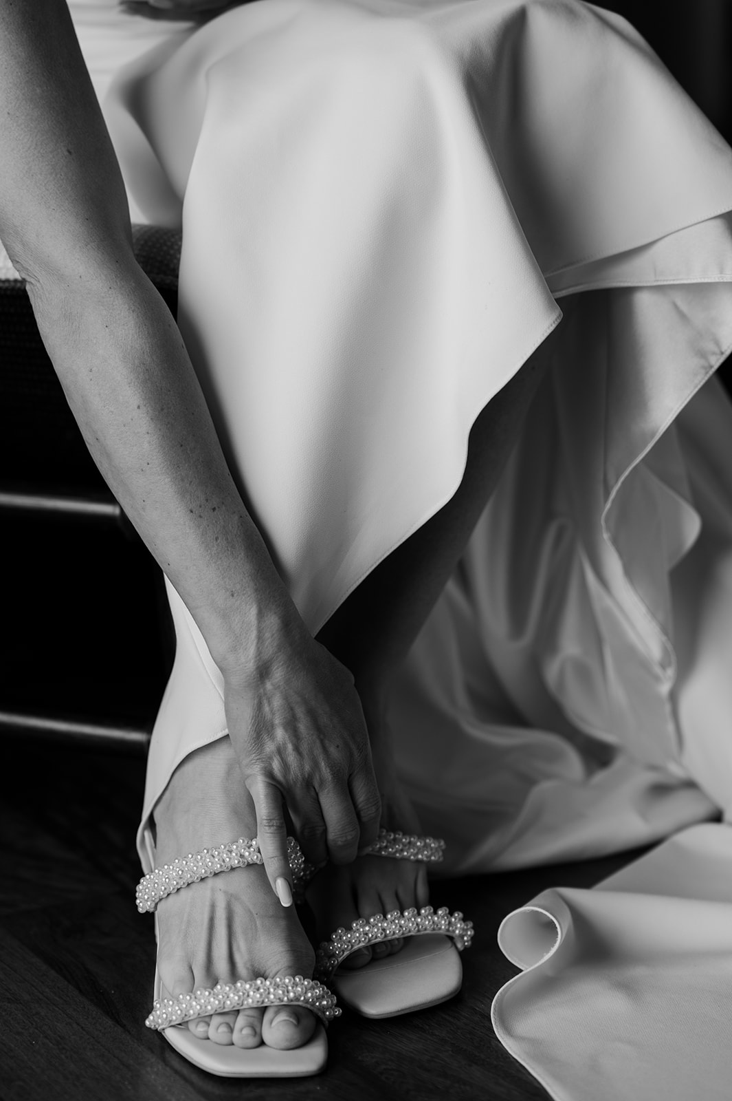 Black and white close-up of the bride putting on her pearl-strap heels with her wedding dress draped beside her.
