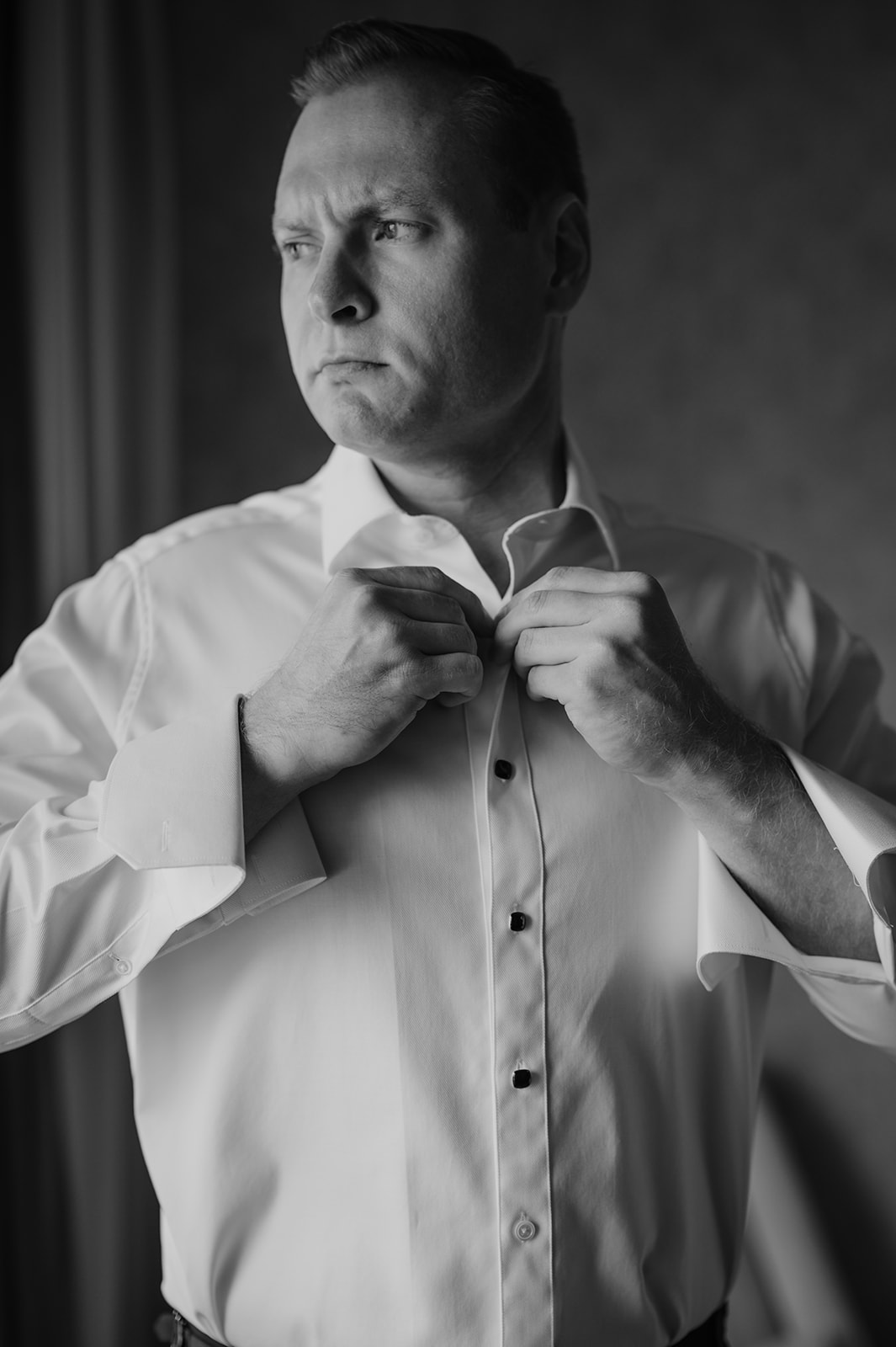 Black and white portrait of the groom buttoning his dress shirt near a window in soft light.
