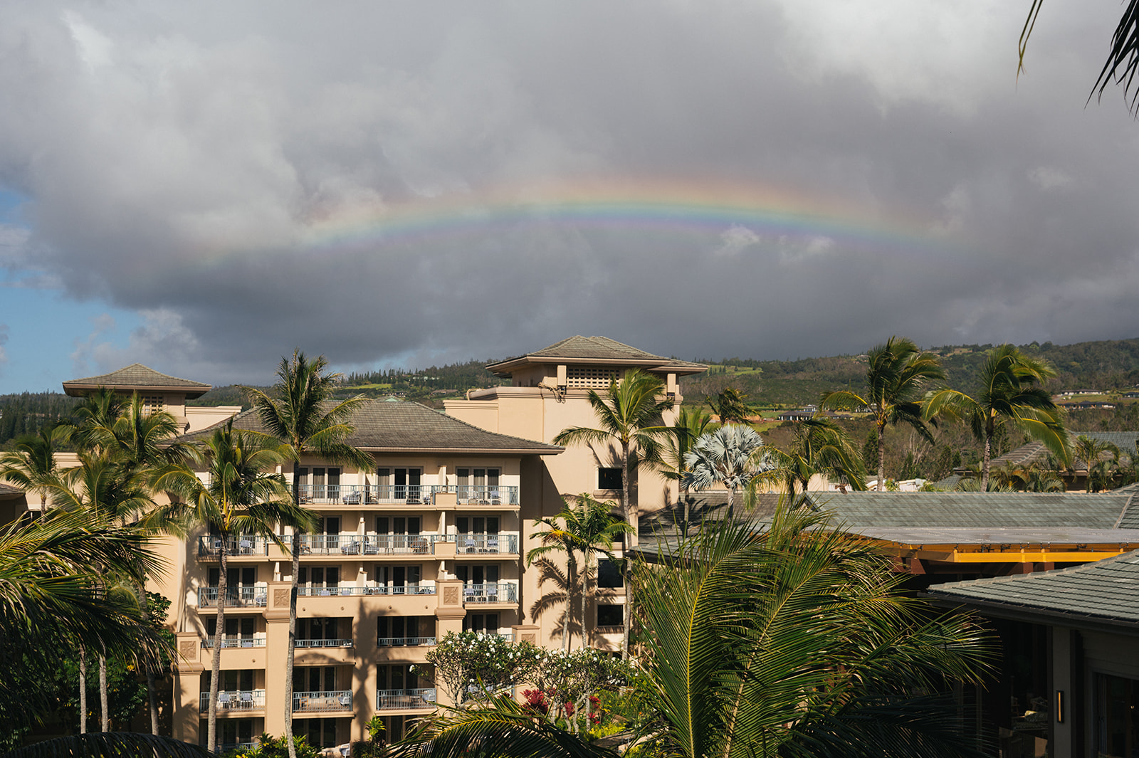 Rainbow arching over the Four Seasons Maui resort surrounded by palm trees and tropical hills.
