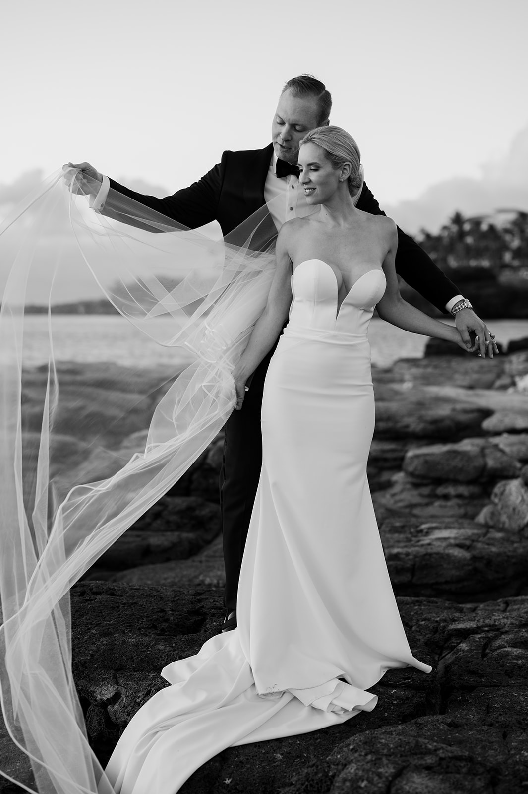 Black and white portrait of the groom lifting the bride’s veil as they stand together on the lava rocks along the Kapalua coastline at sunset.

