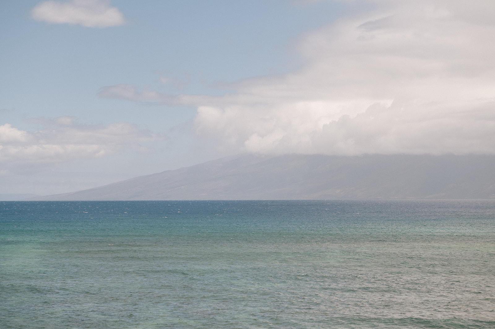Wide view of the Pacific Ocean with the island of Lanai in the distance under soft clouds.
