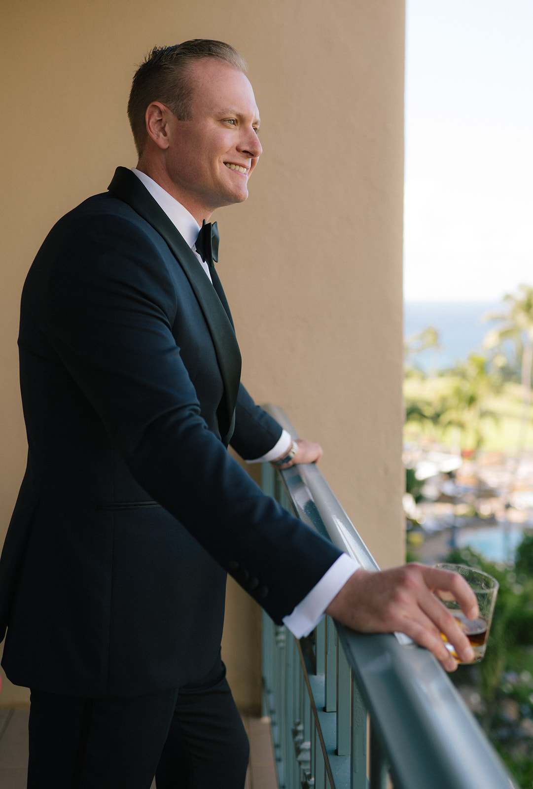 Side profile of the groom standing on a balcony in a tuxedo, overlooking the Maui coastline.
