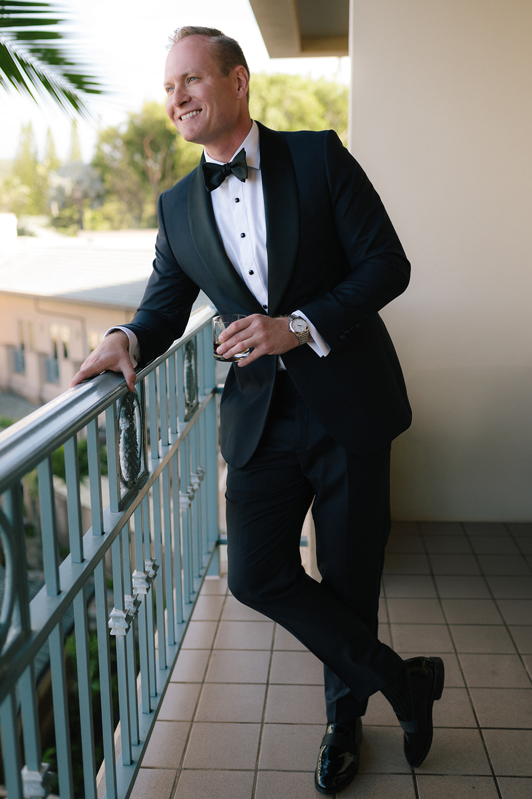 Groom leaning on a balcony railing in a tuxedo at the Four Seasons Maui, smiling in natural light.
