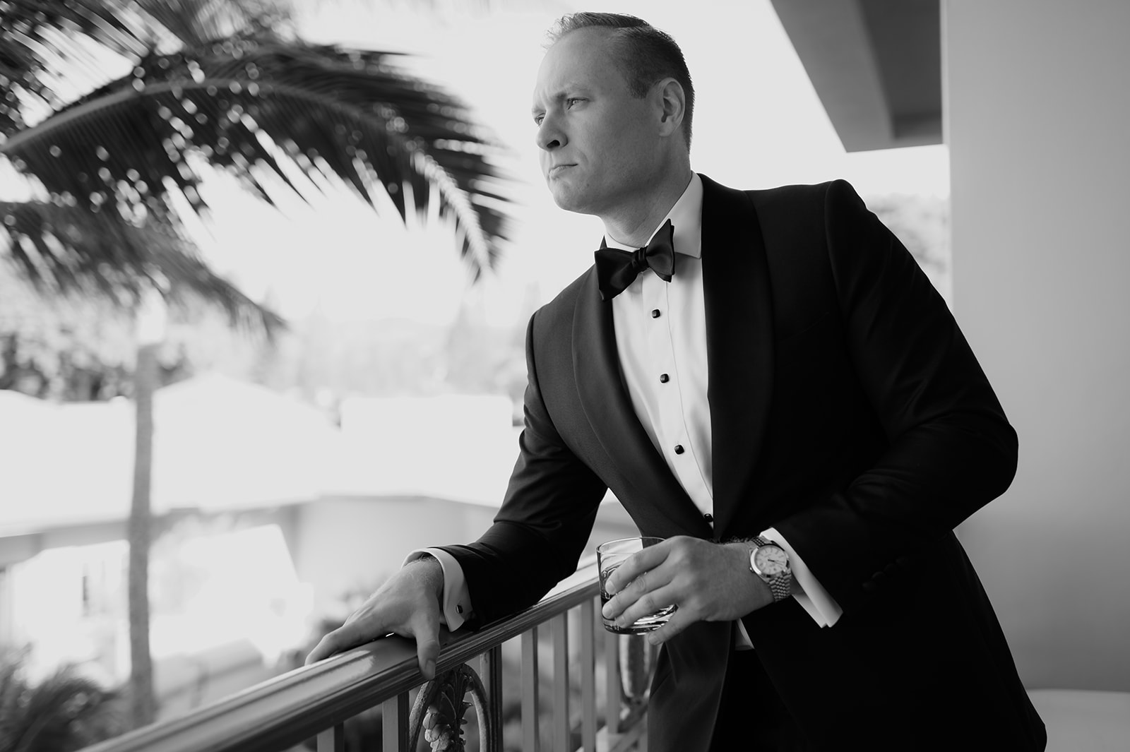 Black and white portrait of the groom in a tuxedo holding a drink and looking out over palm trees in Maui.
