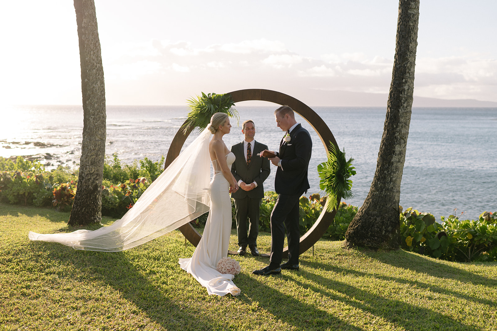 Wide view of a Maui elopement at Merriman’s Kapalua, with the couple and officiant standing beneath a circular arch framed by palm trees and the ocean at golden hour.
