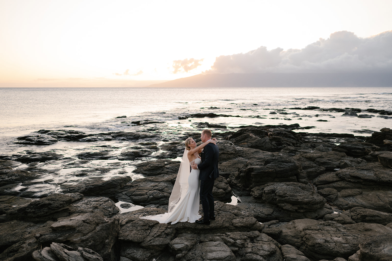 Wide view of the bride and groom embracing on the lava rocks with the Pacific Ocean and sunset sky in Kapalua, Maui.

