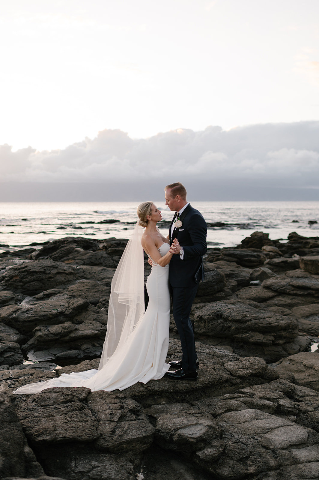 Wide portrait of the bride and groom standing together on the lava rocks with the Pacific Ocean behind them at sunset in Kapalua.
