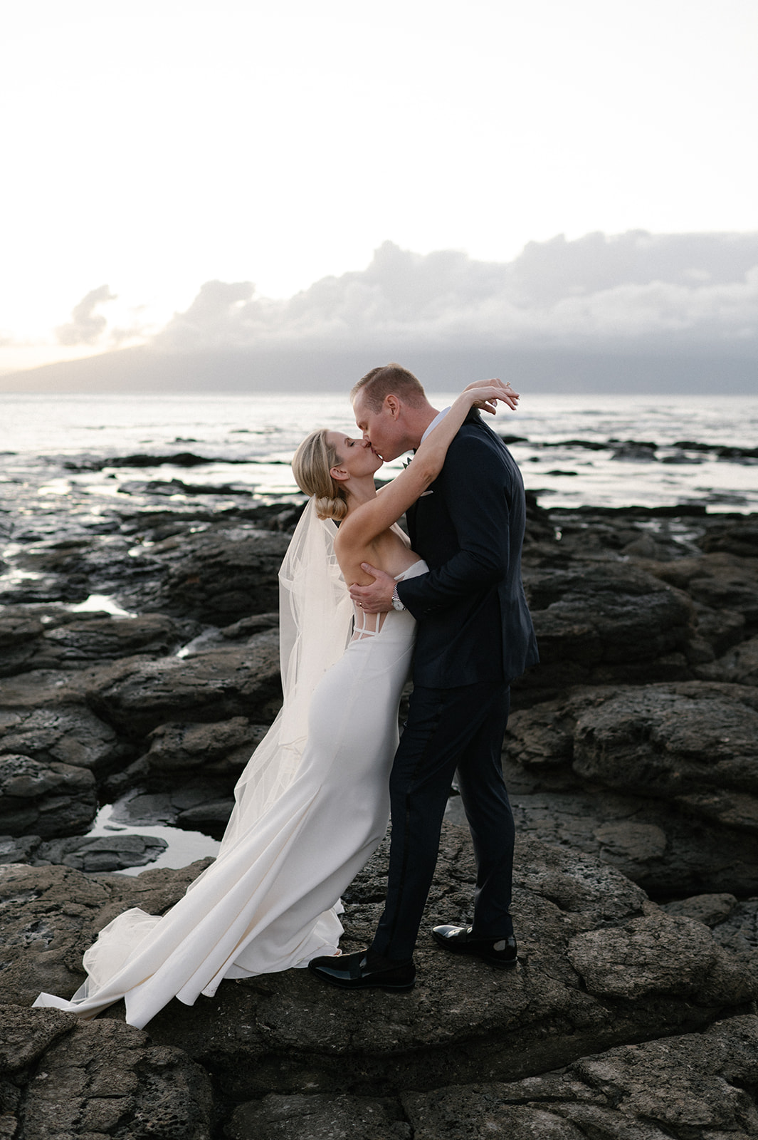 Bride and groom kissing on the lava rocks with the Pacific Ocean behind them during sunset in Kapalua.
